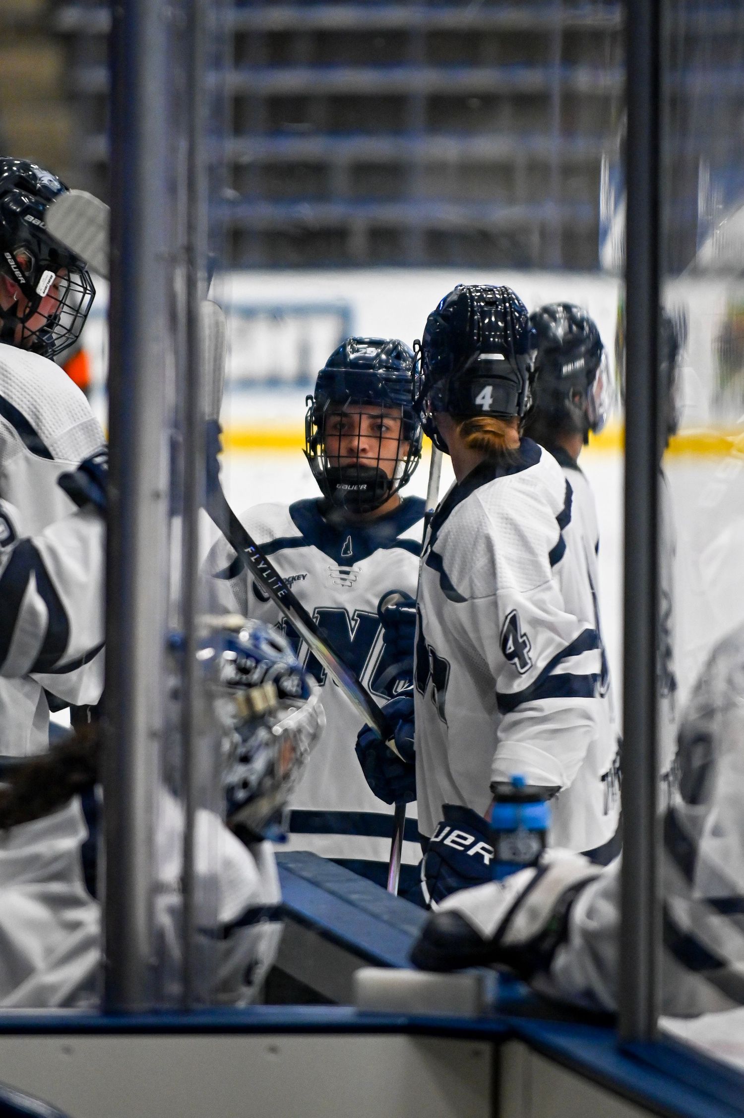 Hockey players sit on the bench during a game wearing white jerseys and protective gear.