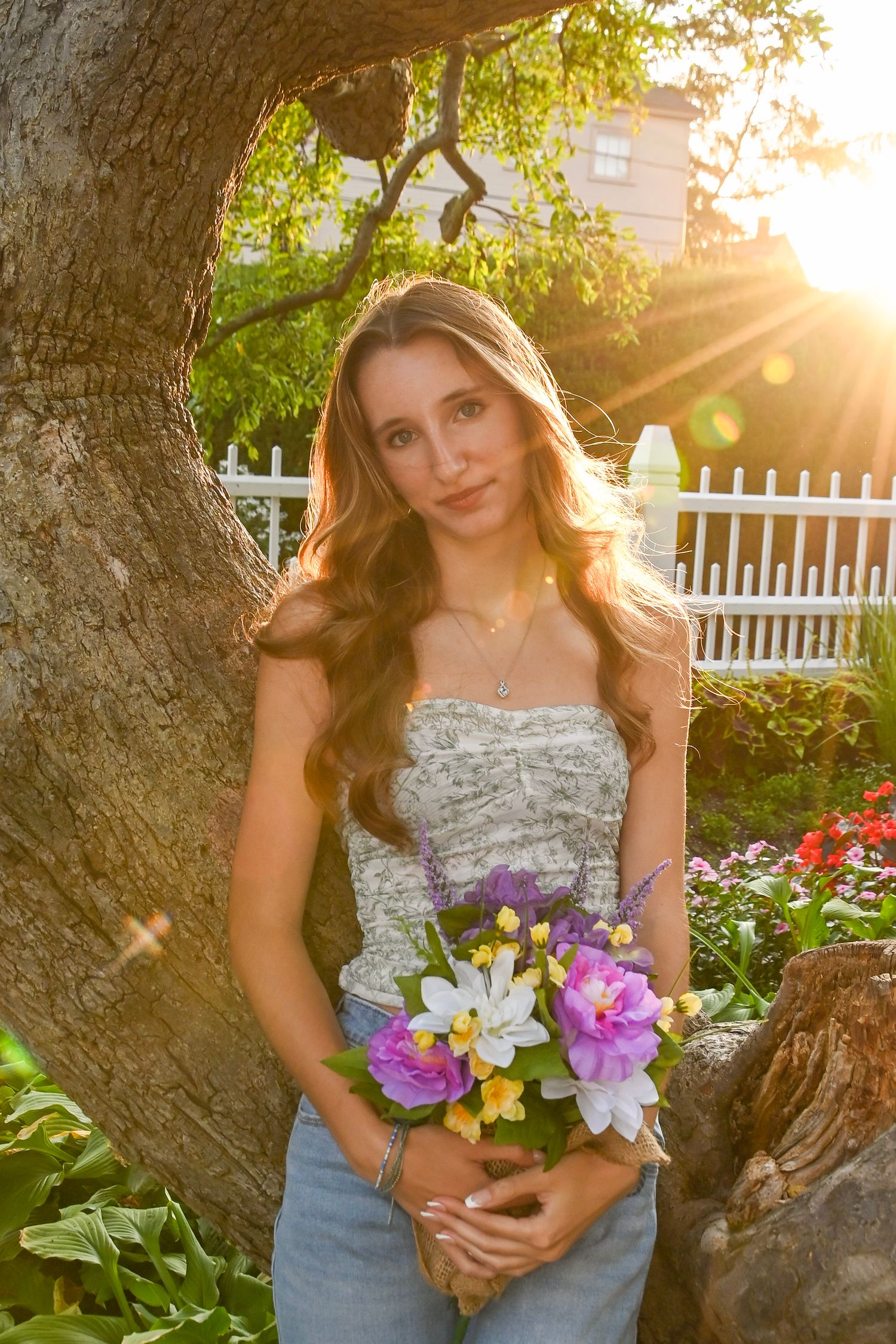A stylish person in a floral bustier and jeans holds colorful flowers while standing by a tree at golden hour.