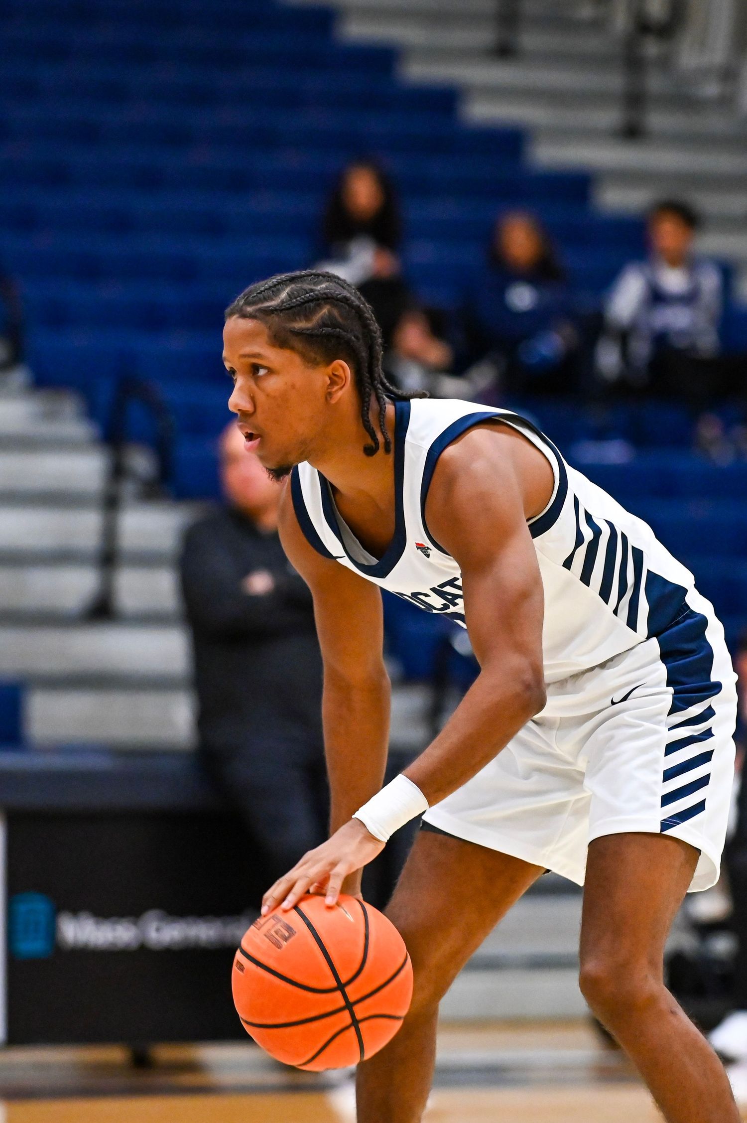 Basketball player in white jersey dribbles ball while crouched in defensive stance during game.