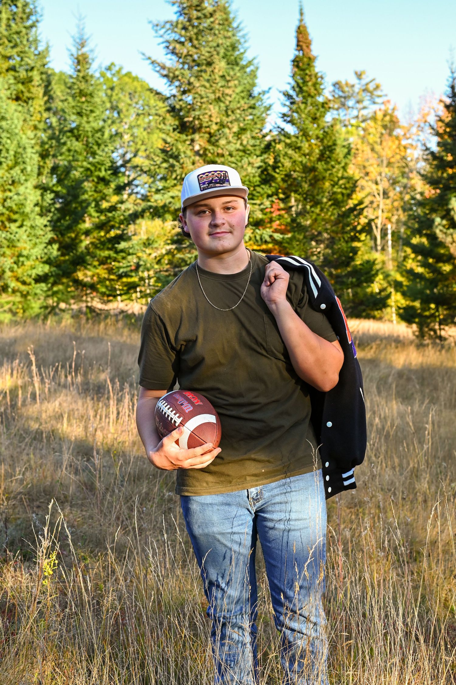 Football player stands in field with varsity jacket and ball against evergreen forest backdrop at sunset.
