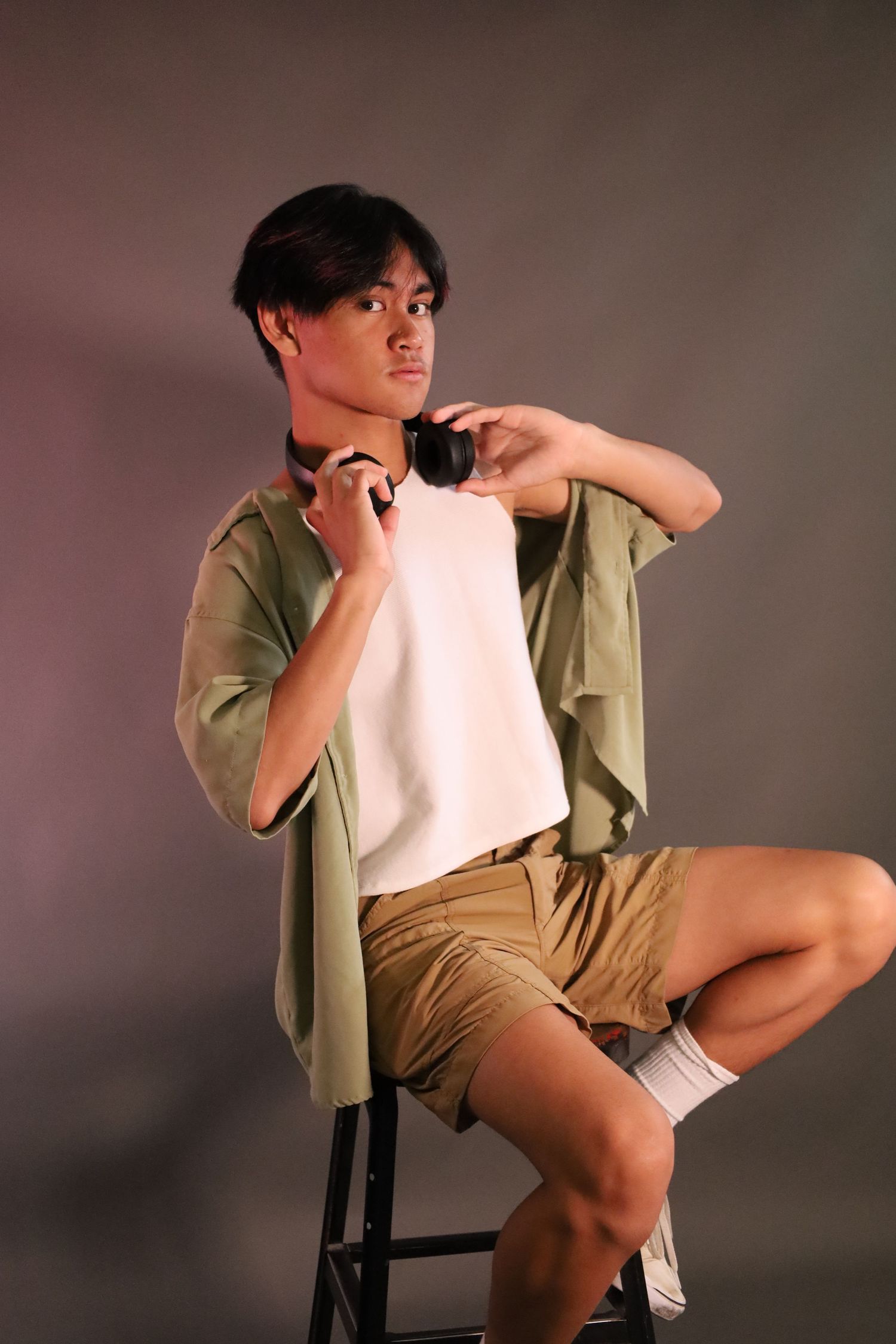 Studio portrait of person in casual summer outfit sitting on stool with headphones.