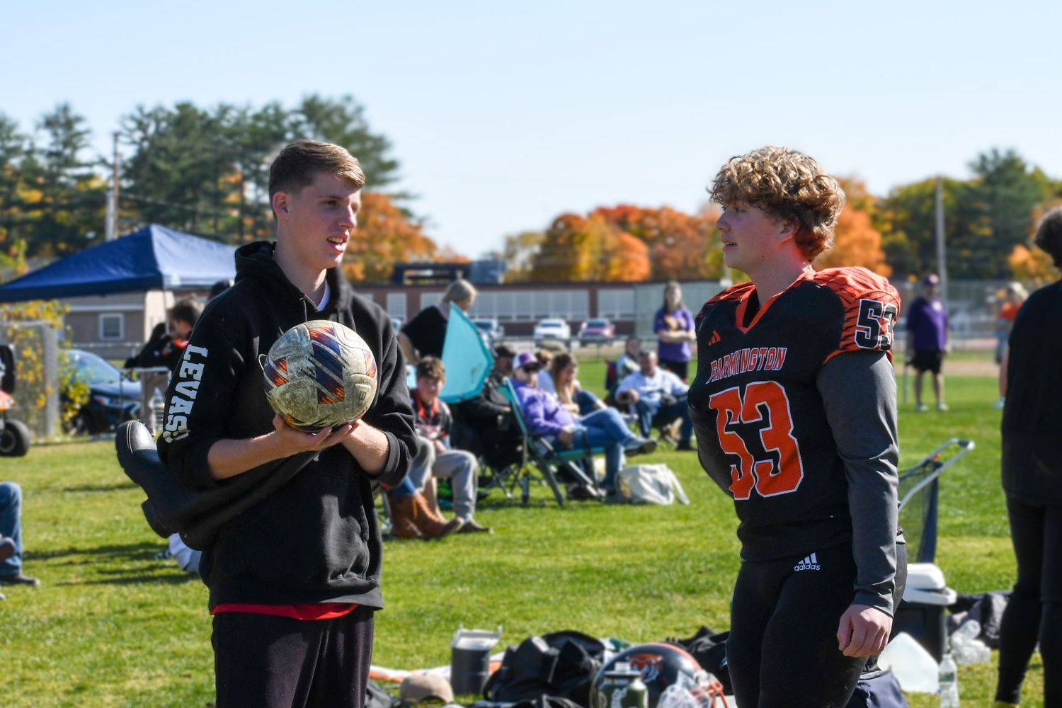 Two lacrosse players talk on a sunny field during a sports event with autumn trees and spectators in background.