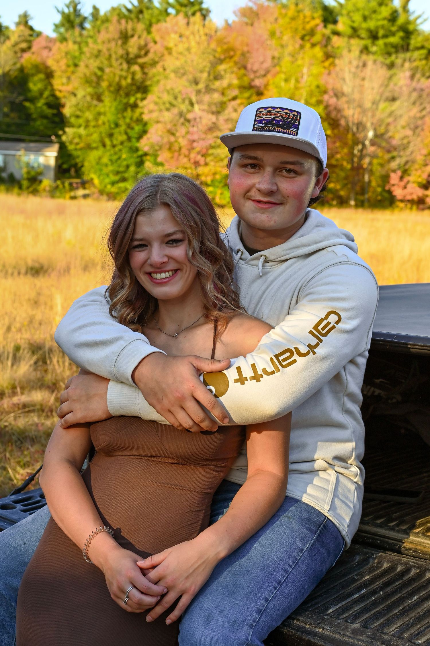 A young couple sitting in the back of a pickup truck, surrounded by autumn foliage in a rural setting.