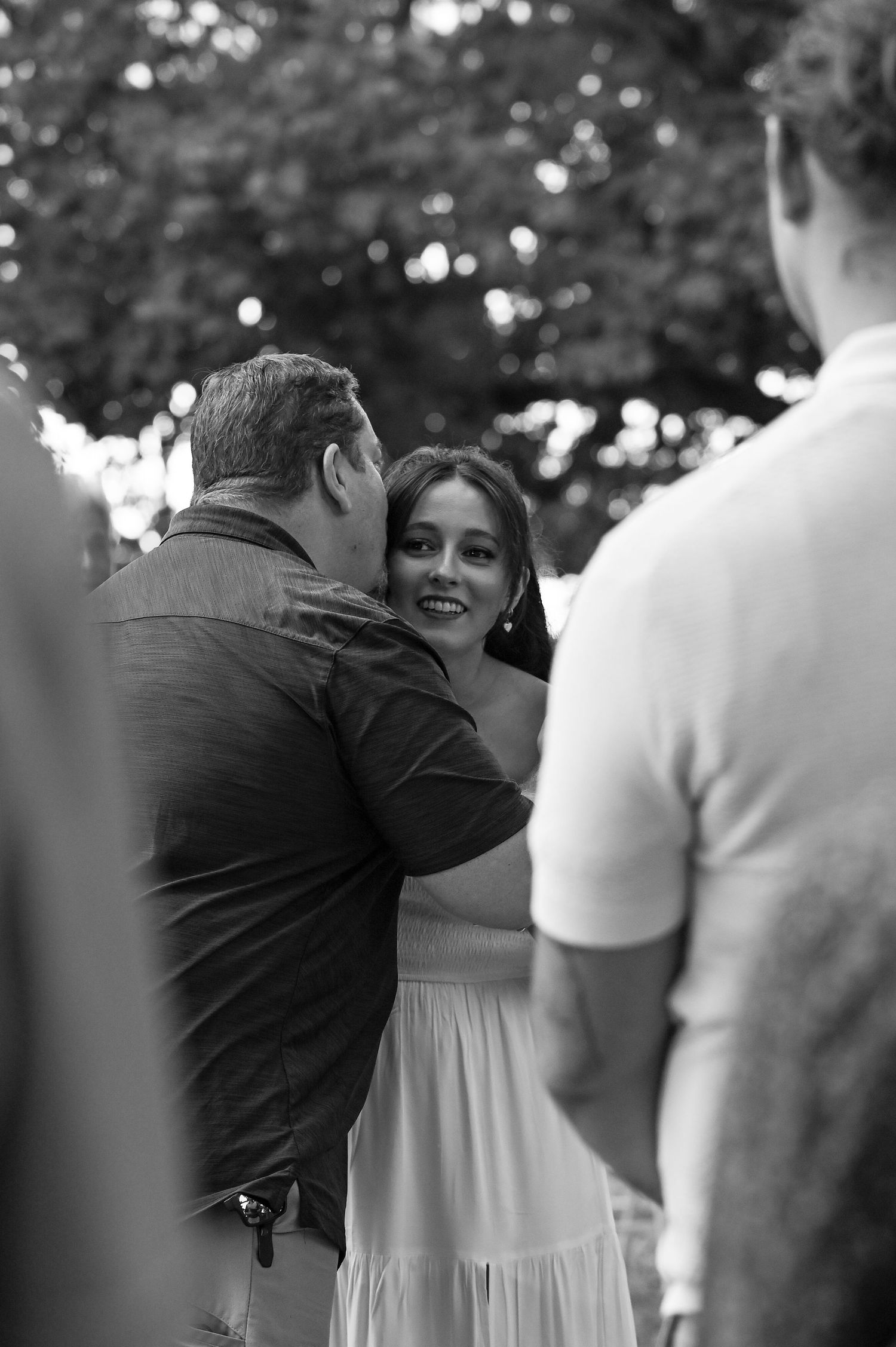 Black and white photo of a smiling woman in a white dress embracing a man outdoors at a social gathering.