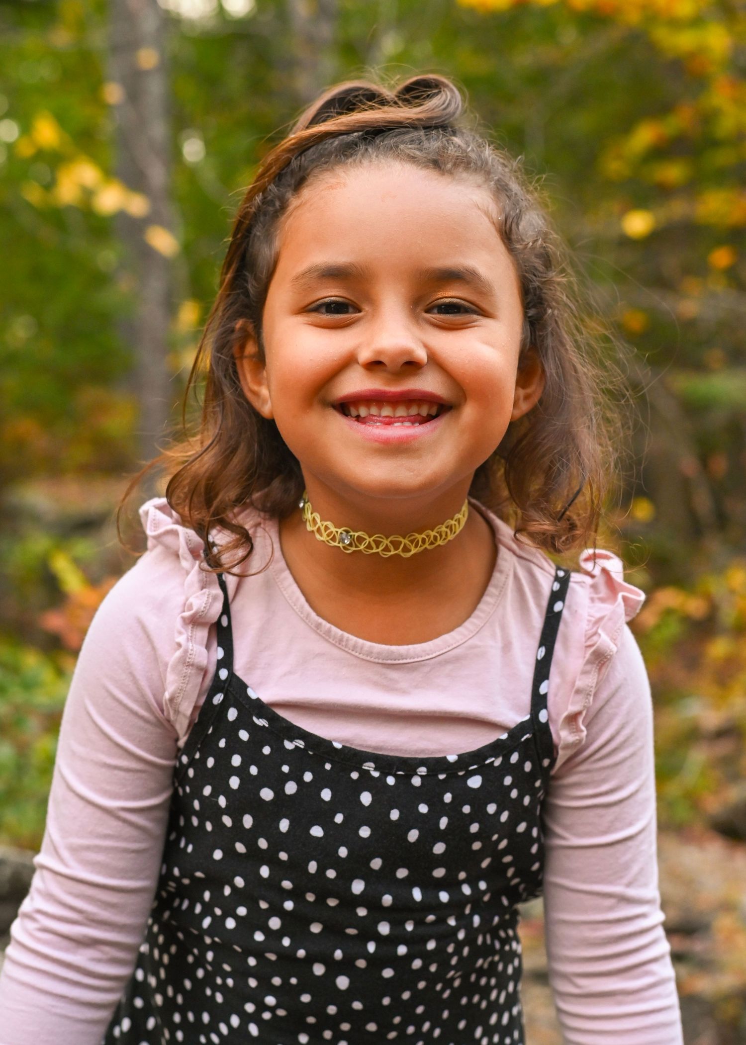 A child in a polka dot dress and pink shirt smiles while playing outdoors among autumn foliage.