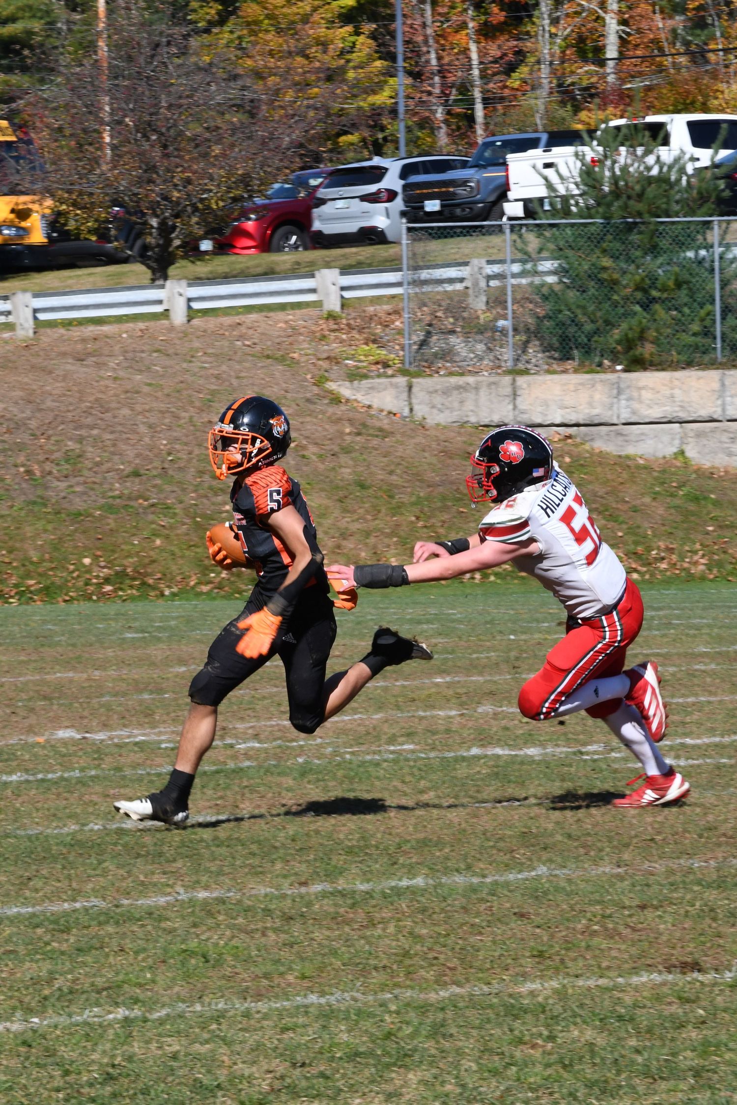 Football player in black uniform runs with ball while opponent attempts tackle on autumn field.
