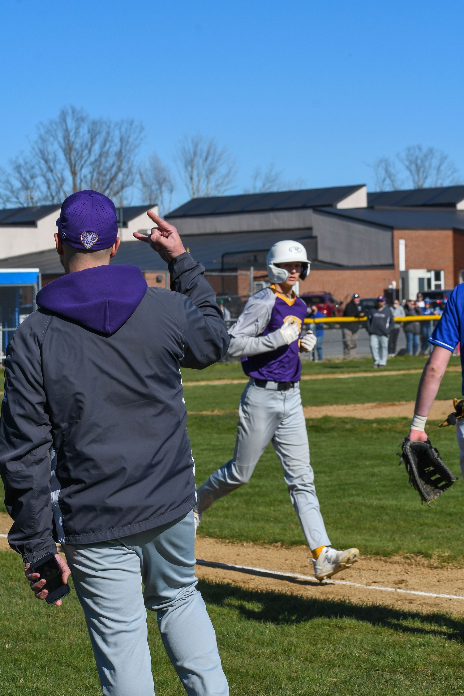 Baseball player in purple uniform runs bases while coach signals from sideline during game.