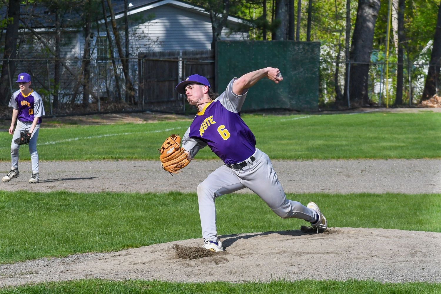 A baseball pitcher in a purple jersey mid-throw on a dirt mound during a game at a grass field.