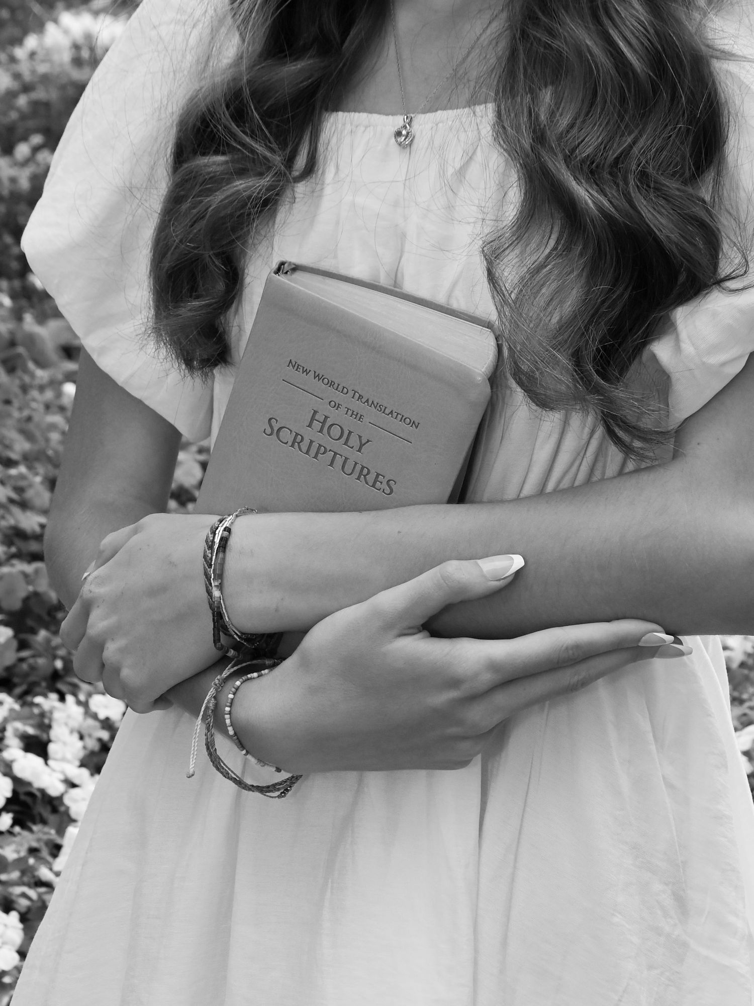 A person in a white dress holds a Holy Scriptures book while wearing multiple bracelets in this black and white photo.