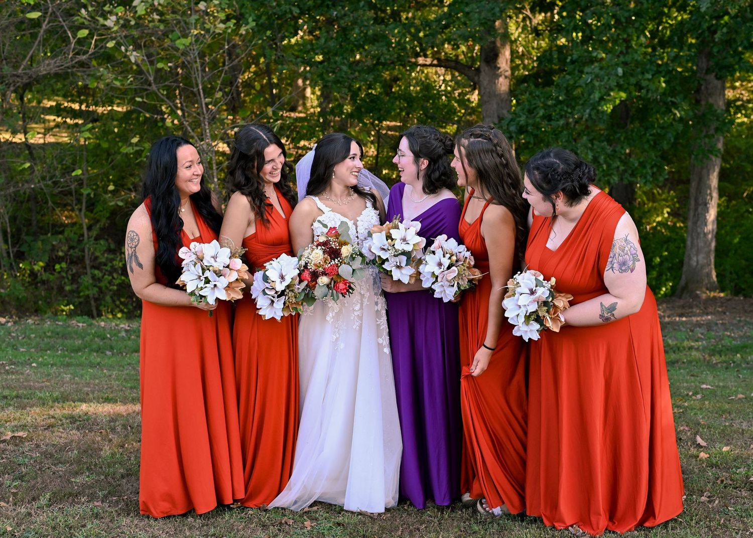 A wedding party in burnt orange and purple dresses stands together holding floral bouquets in an outdoor setting.