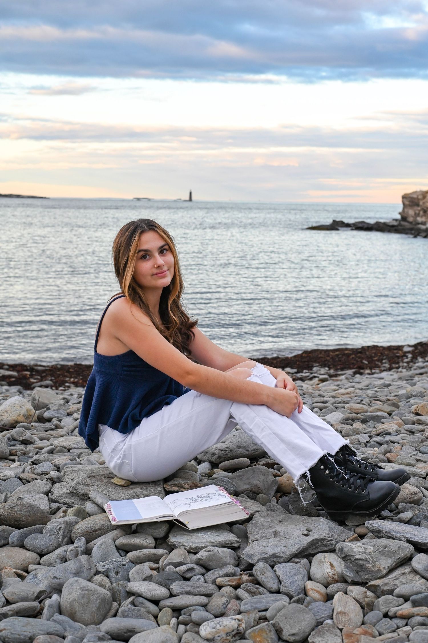 Scenic coastal photoshoot with someone in white pants and navy top sitting on rocky beach at sunset.