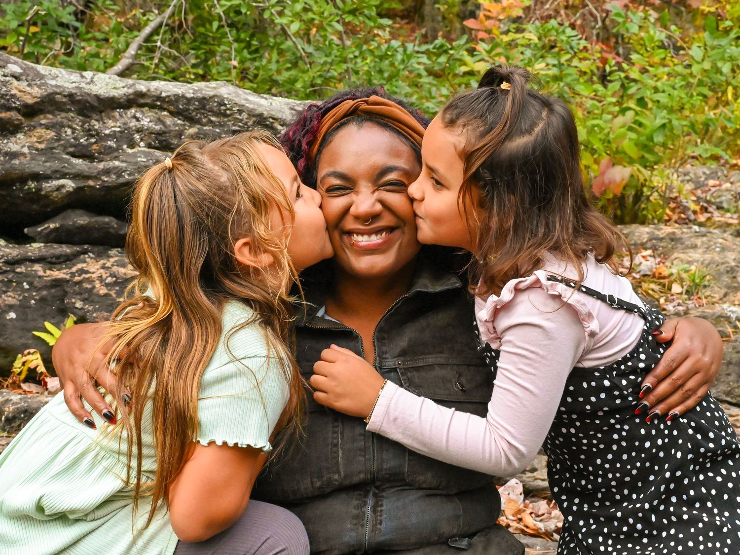 Three people share a joyful moment outdoors while sitting on rocks surrounded by greenery.