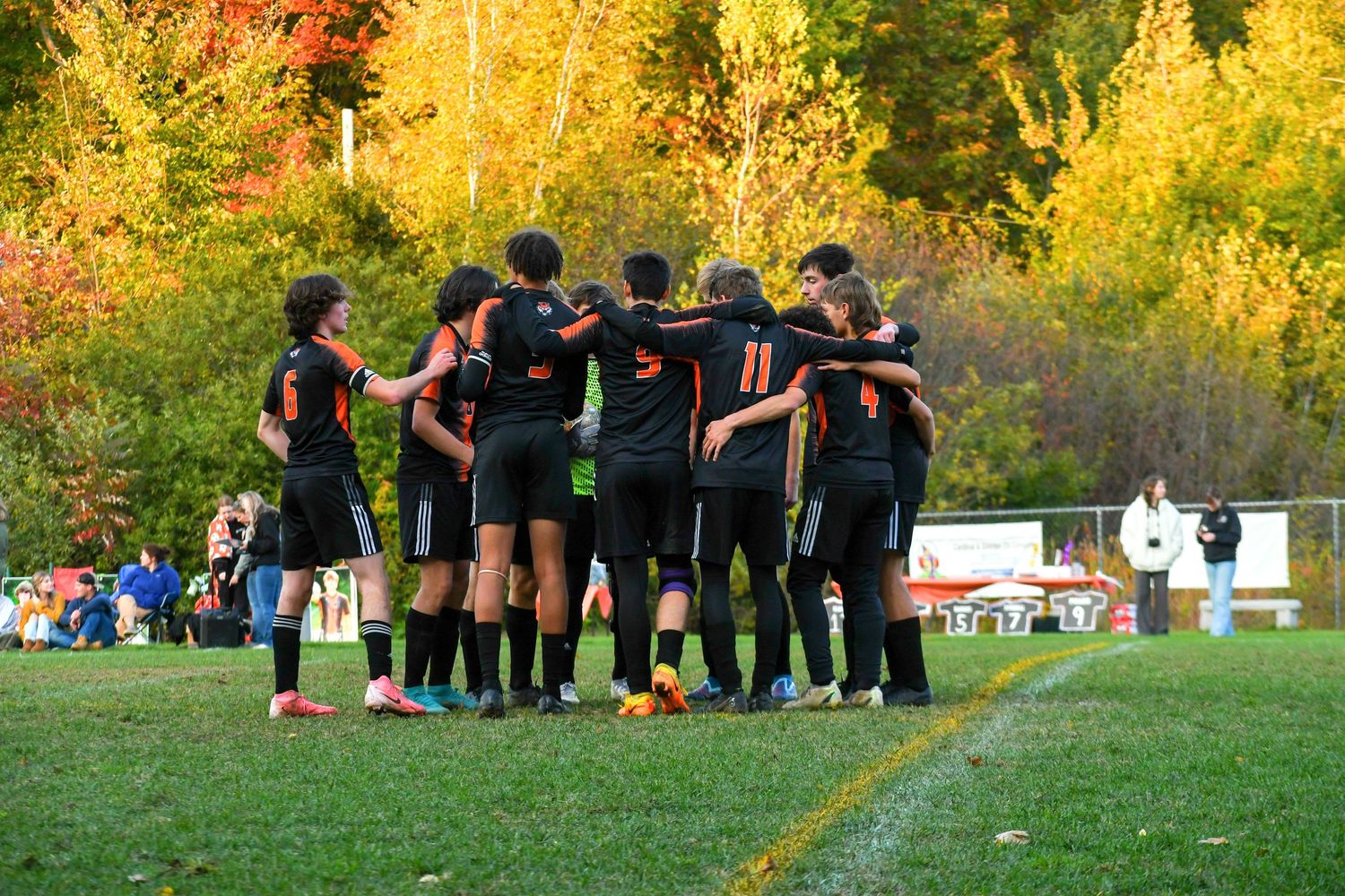 Soccer team huddles together during sunset with fall foliage in background.