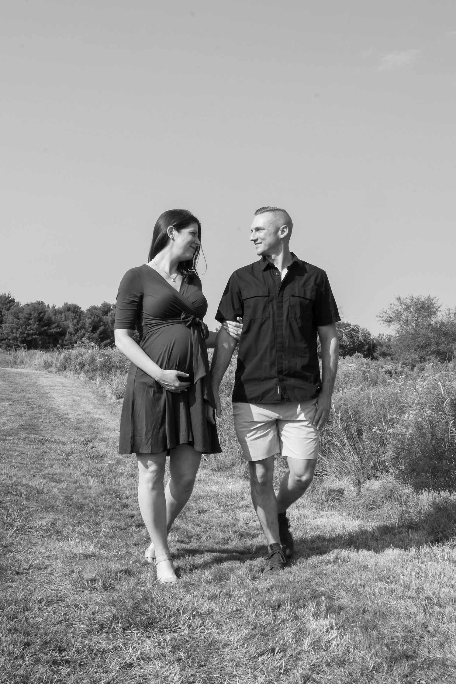 A couple walks together on a rural path, with the woman wearing a dress and the man in casual summer attire.