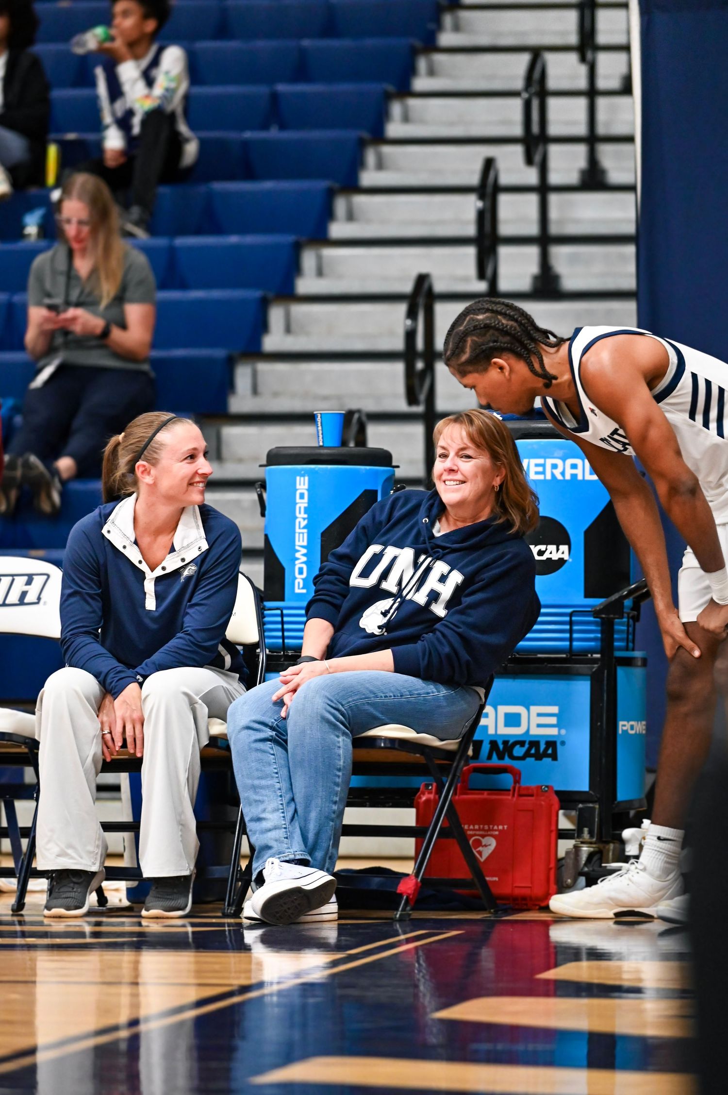 Players and coaches sit on sideline benches during a UNH basketball game in a gymnasium.