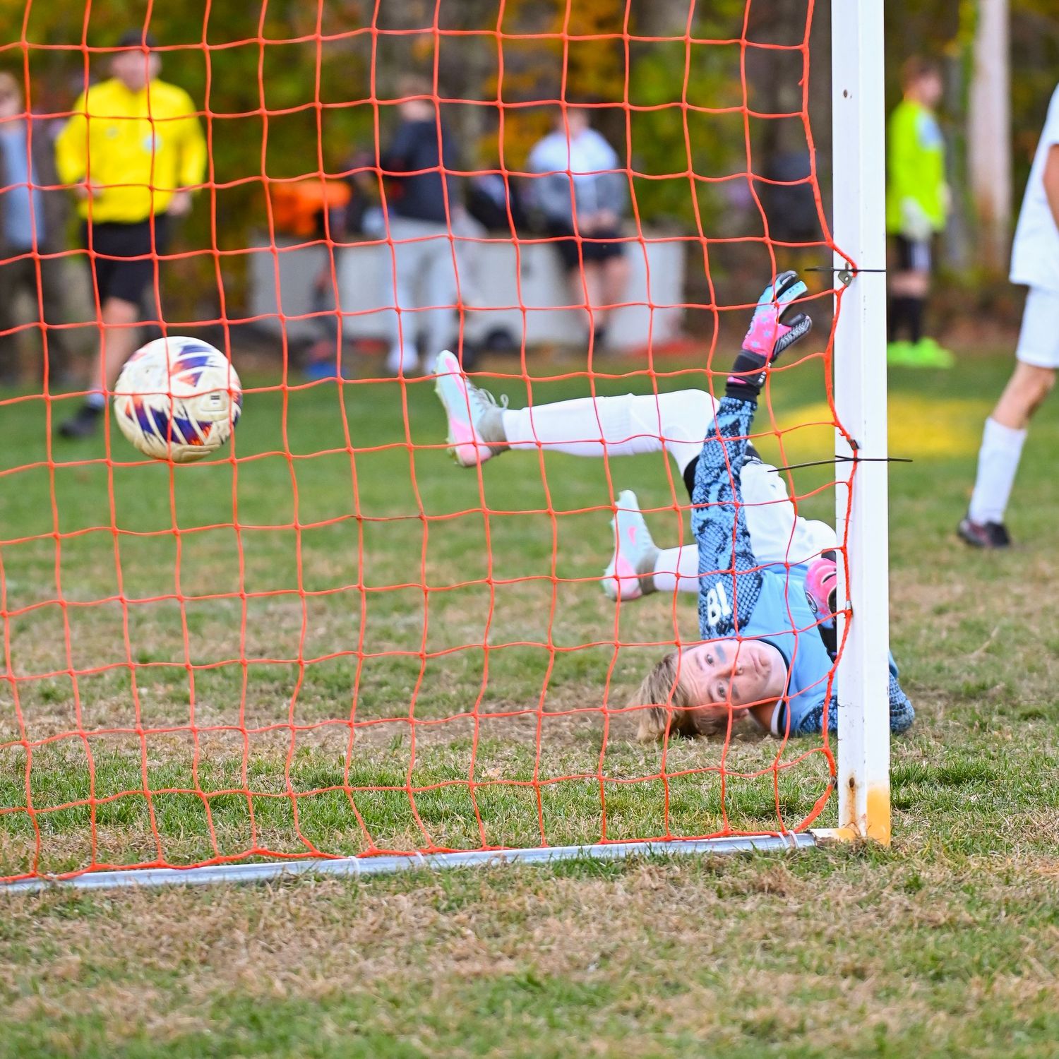 Soccer ball rolling into goal net during match with goalkeeper attempting save on grass field.