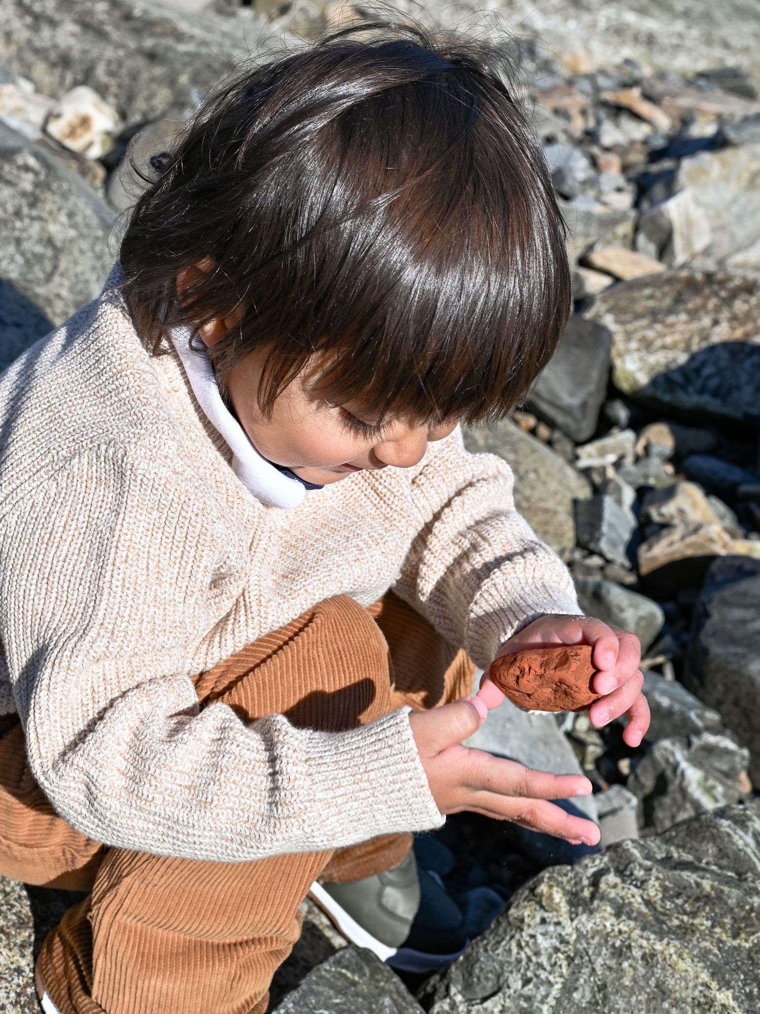 A child in a beige sweater examines rocks and minerals while sitting on a rocky surface outdoors.