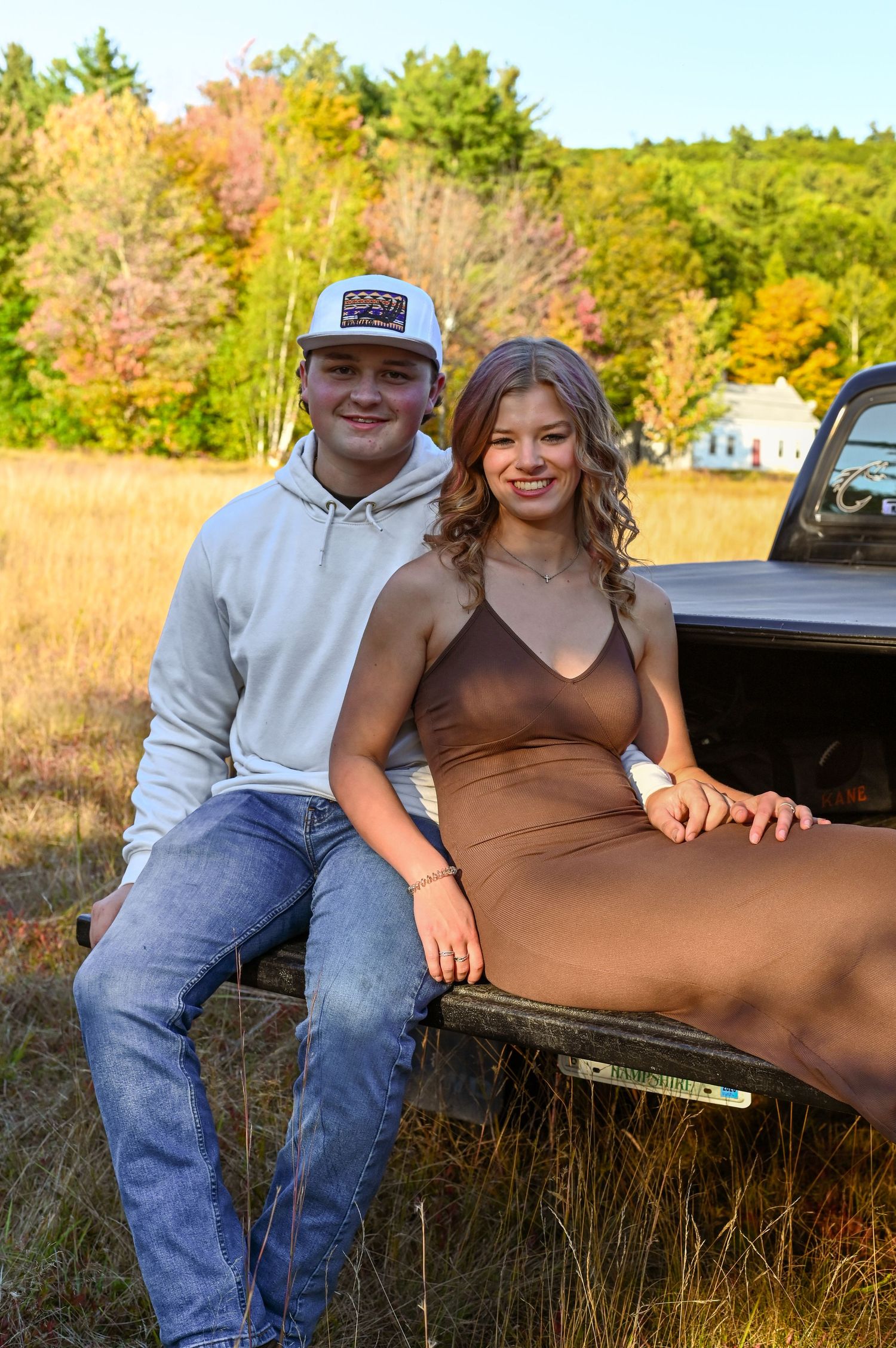 A young couple posing together on the back of an old pickup truck in a scenic, autumn-colored forest.