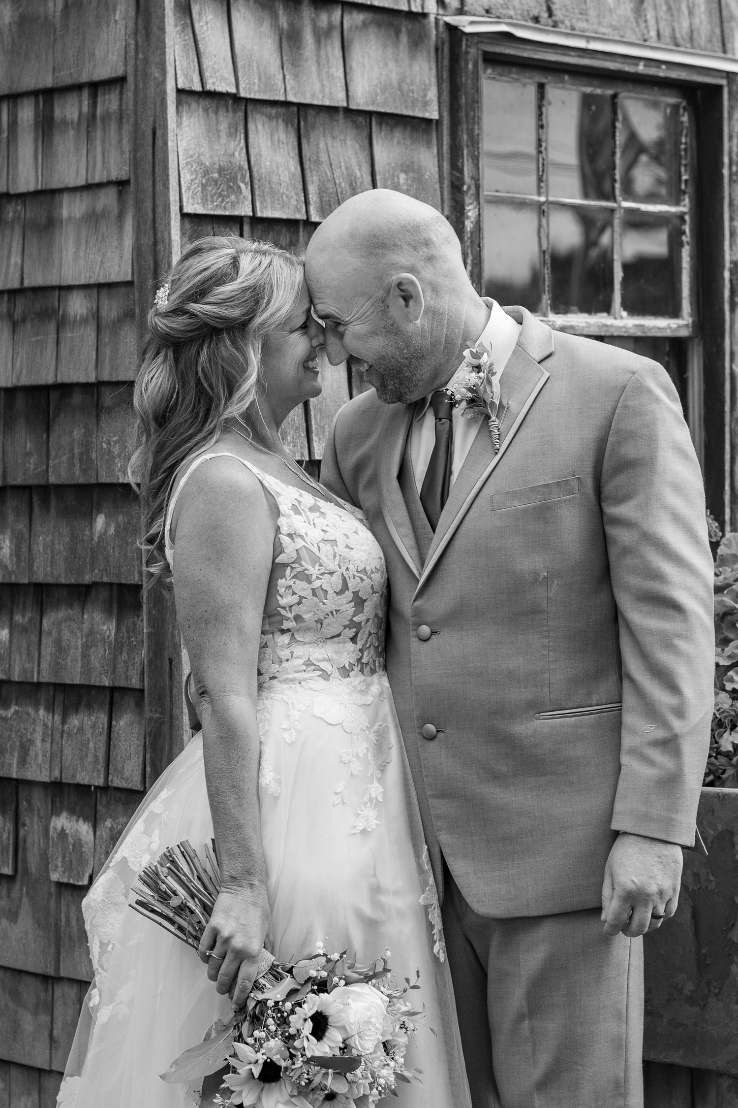 Black and white wedding photo of a bride in a lace gown holding a bouquet touching foreheads with a groom in a suit by a shingled building.