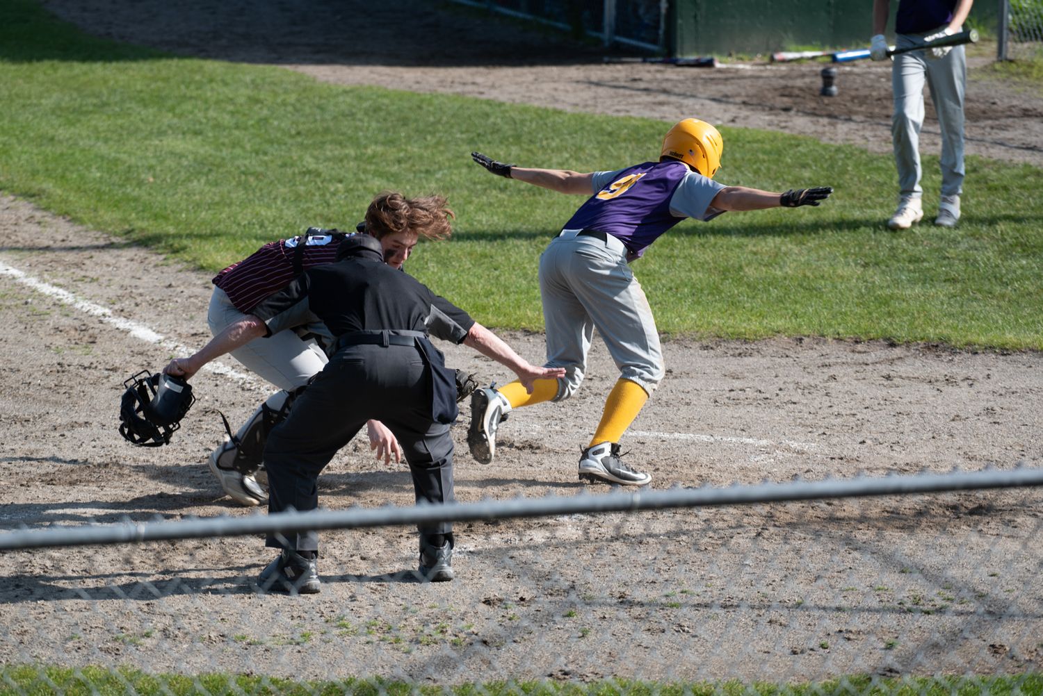 Baseball player in purple uniform swings at ball while catcher and umpire watch at home plate during game.
