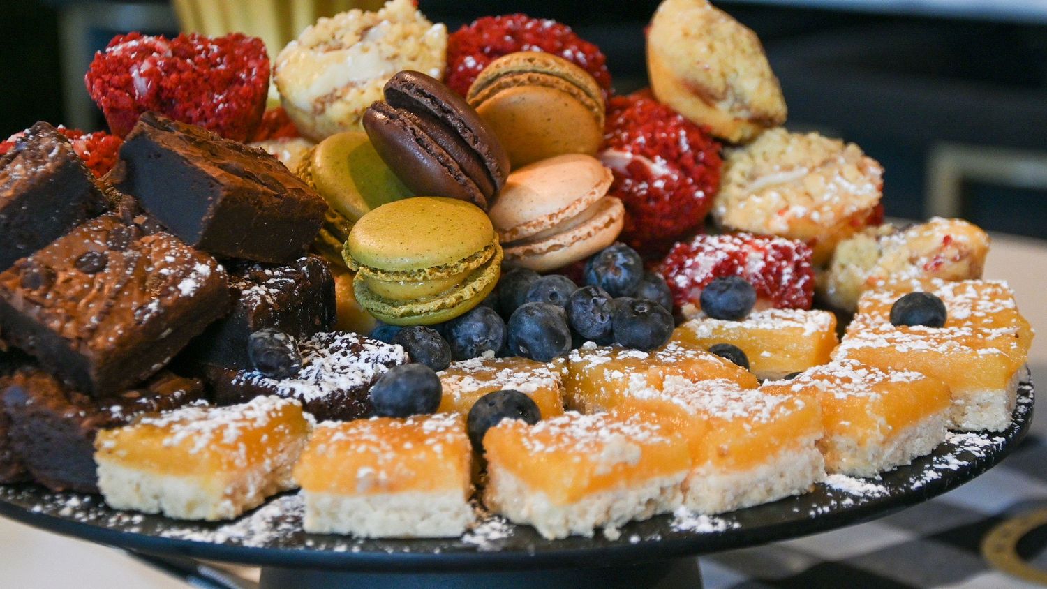 A dessert platter featuring macarons, brownies, blueberries, raspberries and powdered citrus bars arranged on a dark plate.