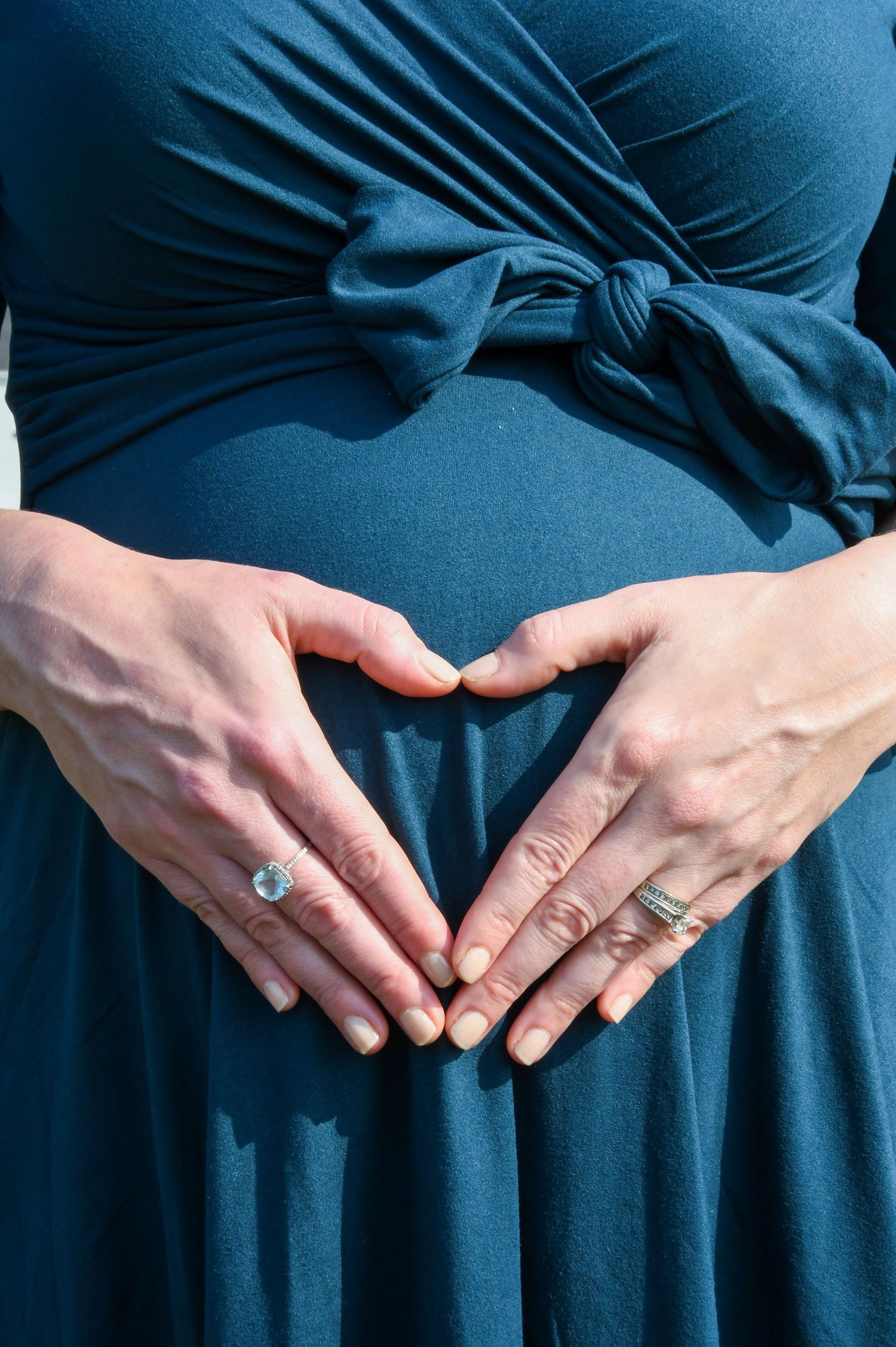 Hands form a heart shape over a pregnant belly wearing a blue maternity dress with rings visible on the fingers.