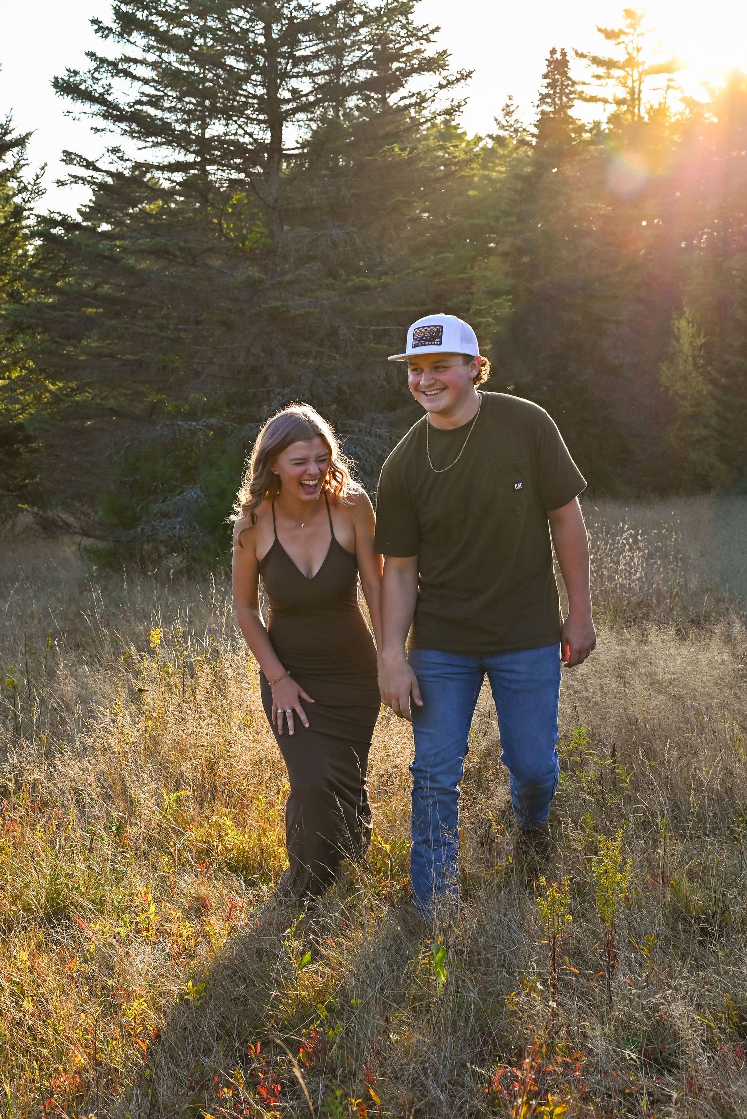 A man and woman walking together in a field at sunset, surrounded by pine trees.