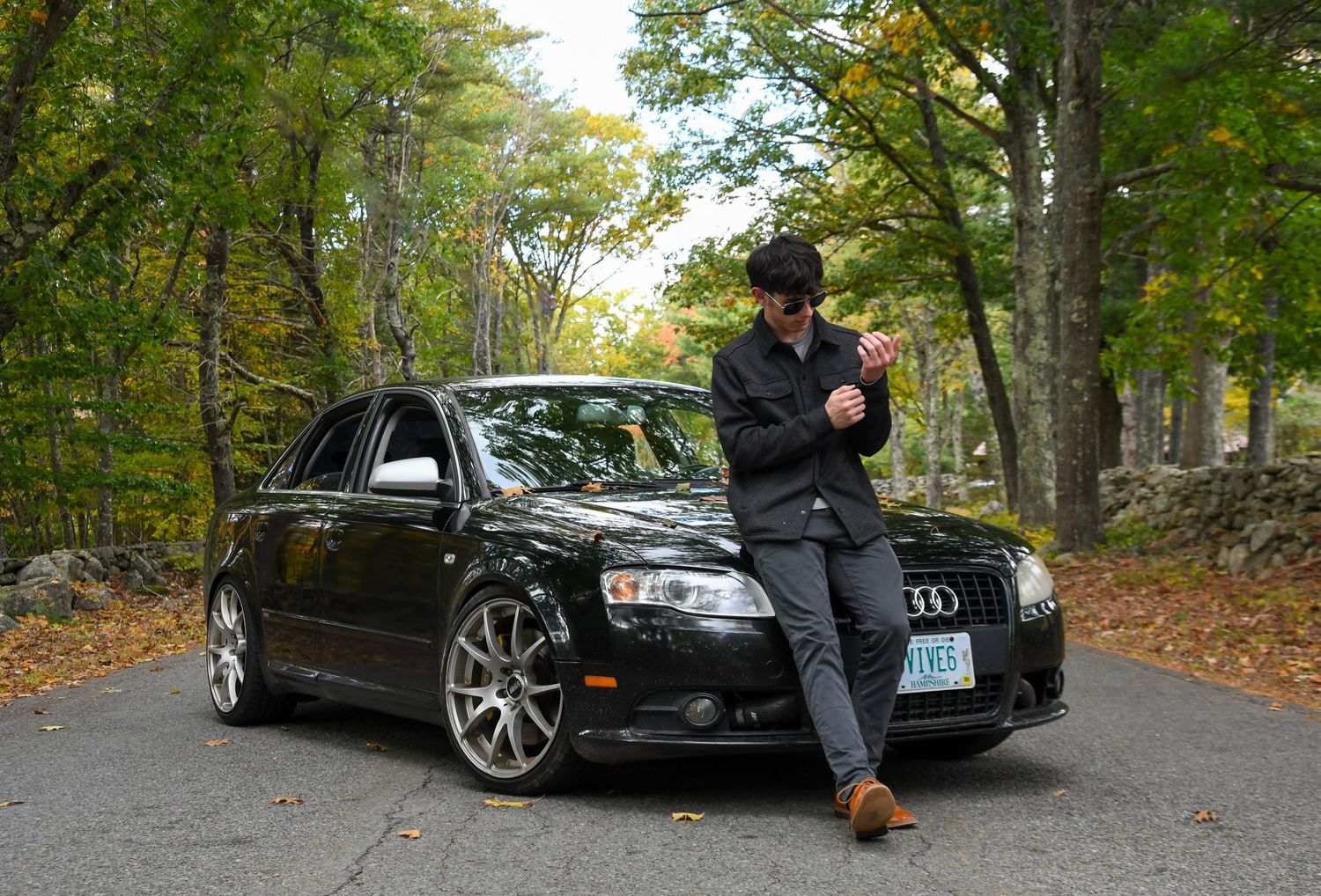 Casual portrait with person leaning against black Audi on tree-lined road.