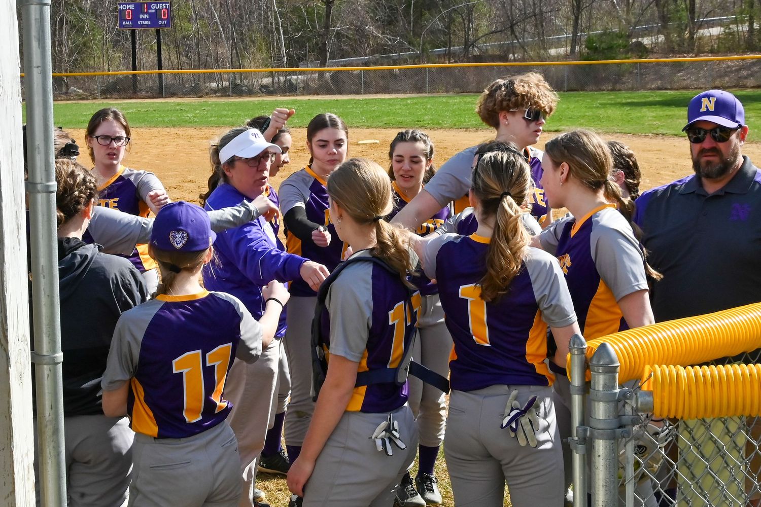 A softball team huddles together on the field wearing purple and white uniforms during a game.