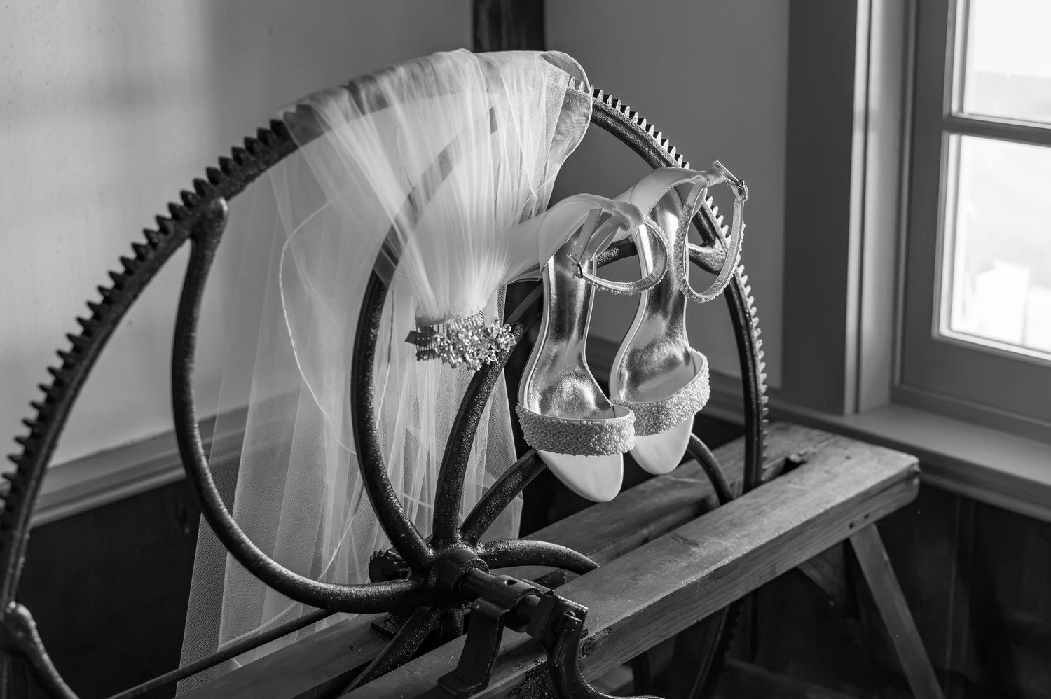 Black and white photo of bridal shoes and a veil with a jeweled hair clip draped over a vintage spinning wheel by a sunlit window.