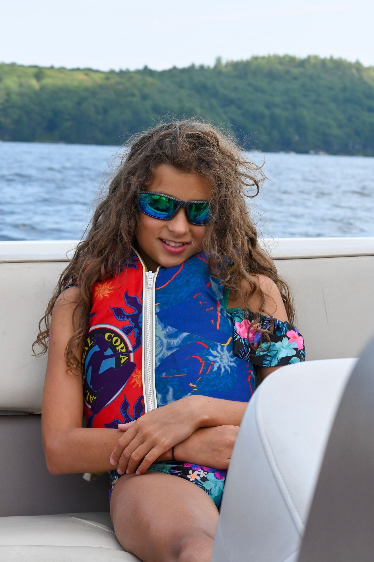 Someone relaxes on a boat wearing a colorful floral swimsuit and blue sunglasses near a scenic shoreline.