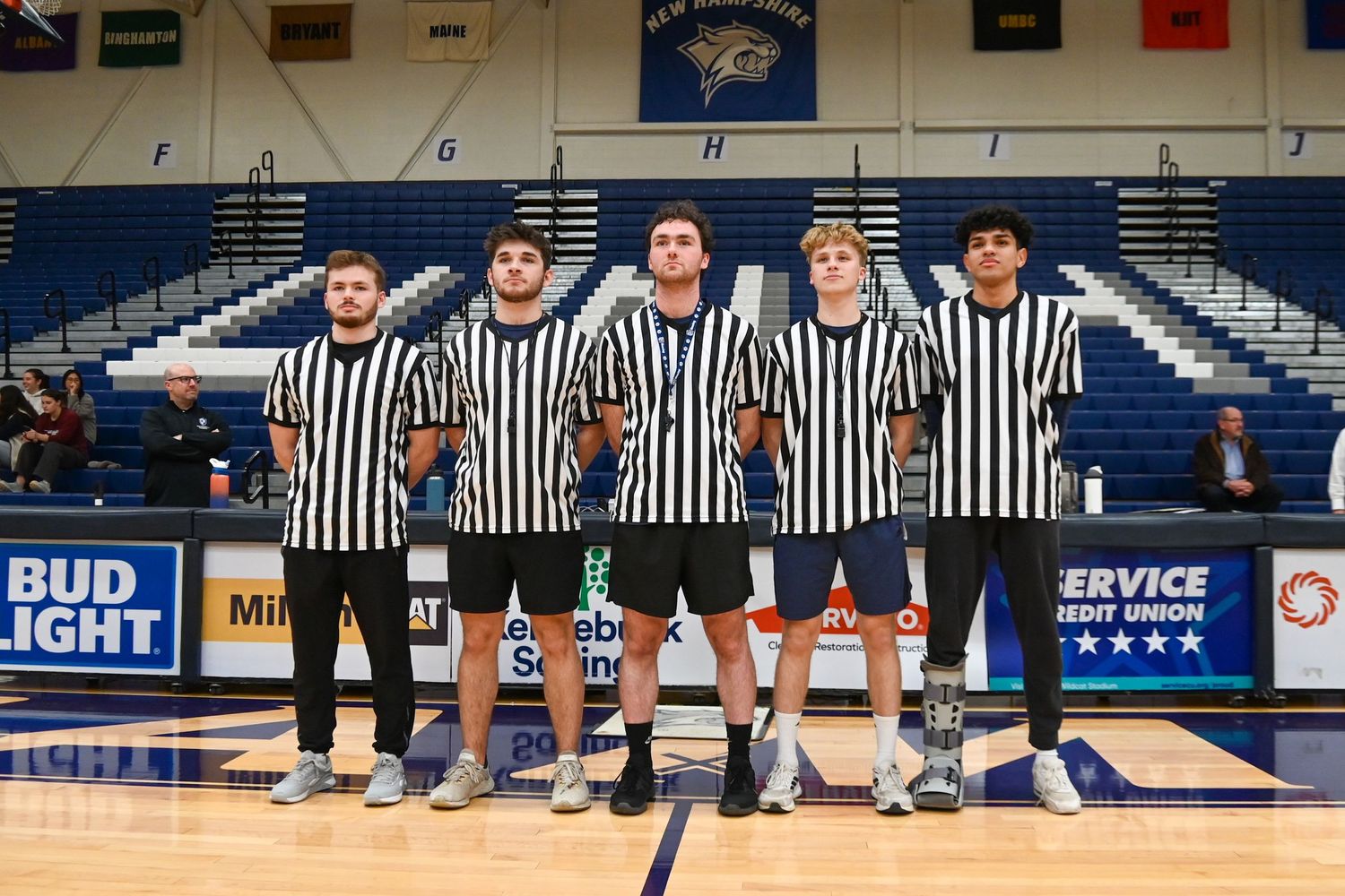 Group of basketball referees in striped uniforms pose together on court under arena lights.