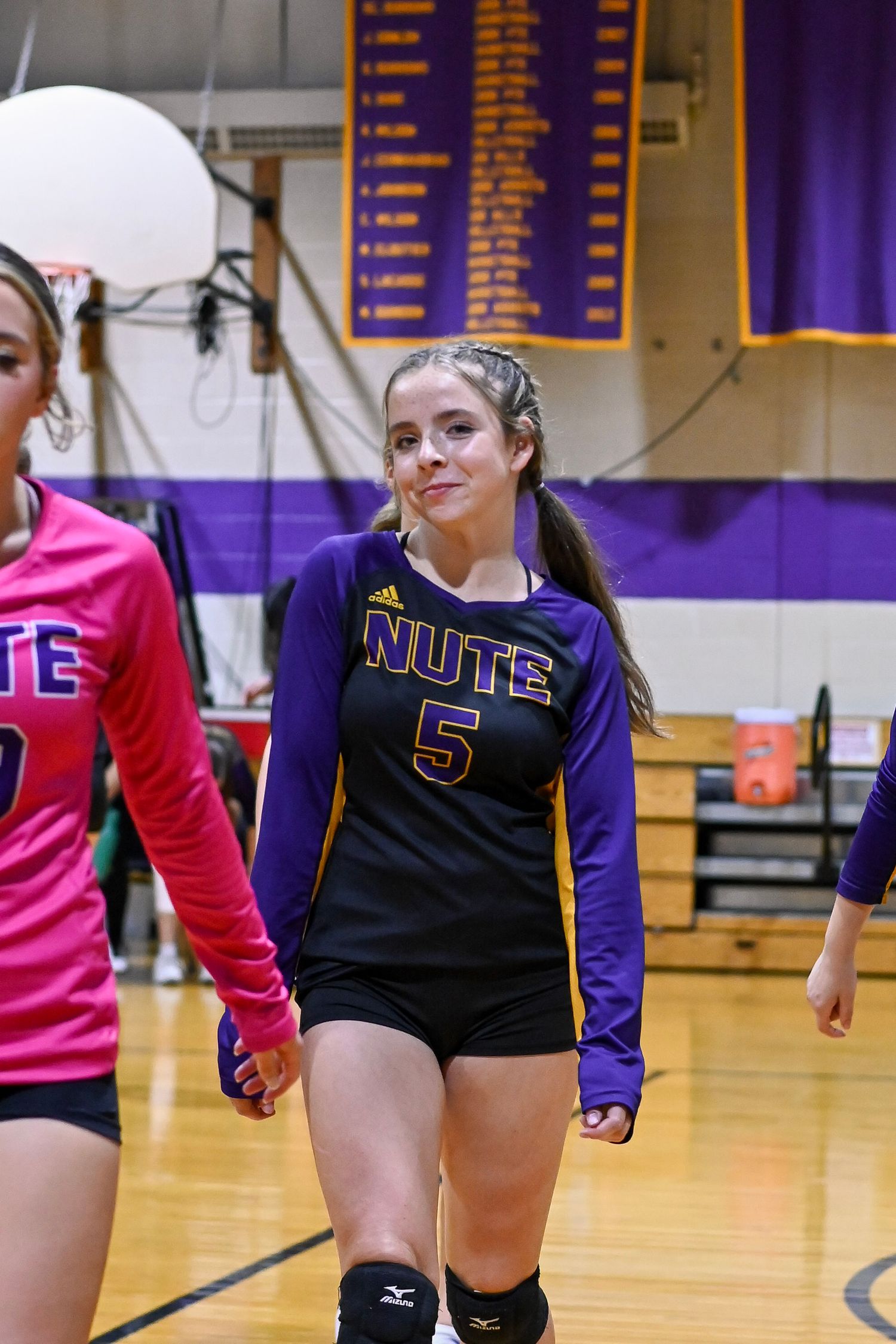 Volleyball players in purple and black uniforms position themselves on a wooden court during a game.