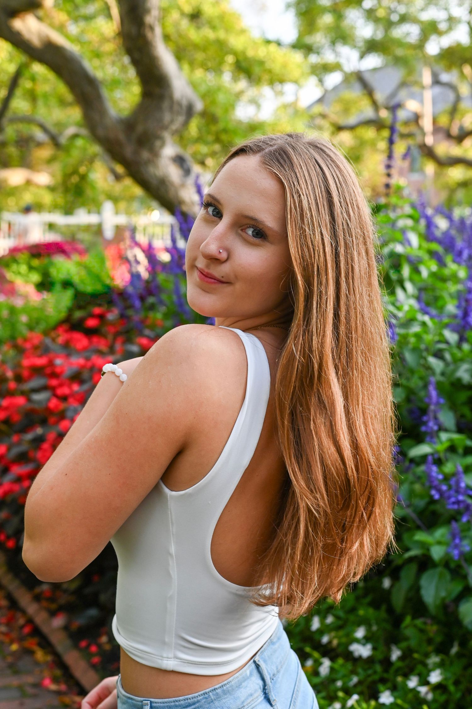 A person in a white tank top poses in a garden with vibrant red and purple flowers on a sunny day.