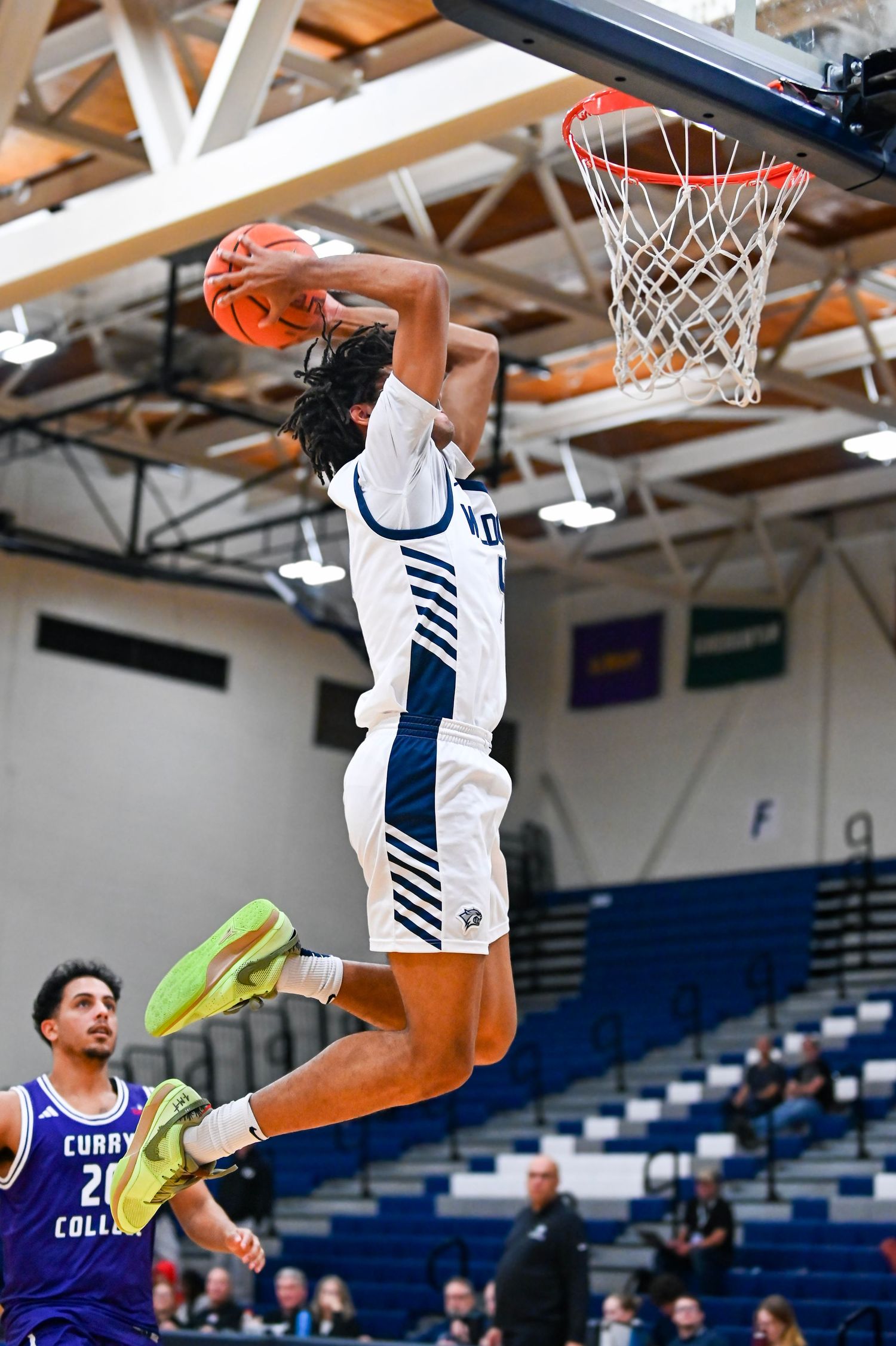 Basketball player in white and navy uniform soars through the air for a powerful slam dunk during indoor game.