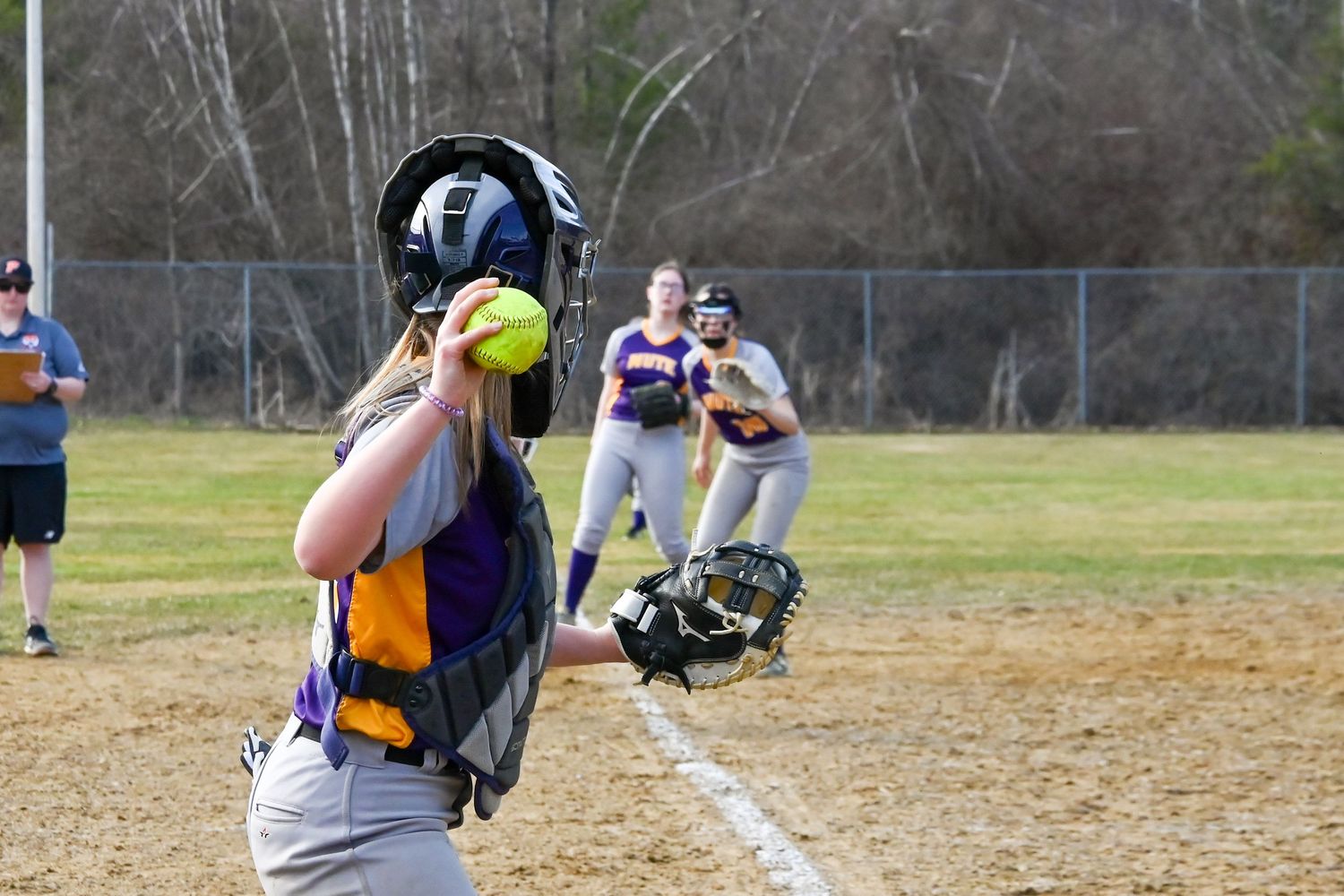 Softball catcher looks up to catch a pop fly during a game on a sunny day while teammates watch in the background.