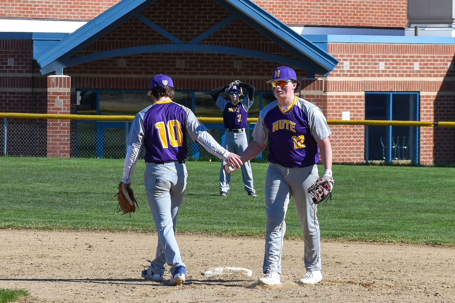 Baseball teammates interact on the field during a game with brick building in background.