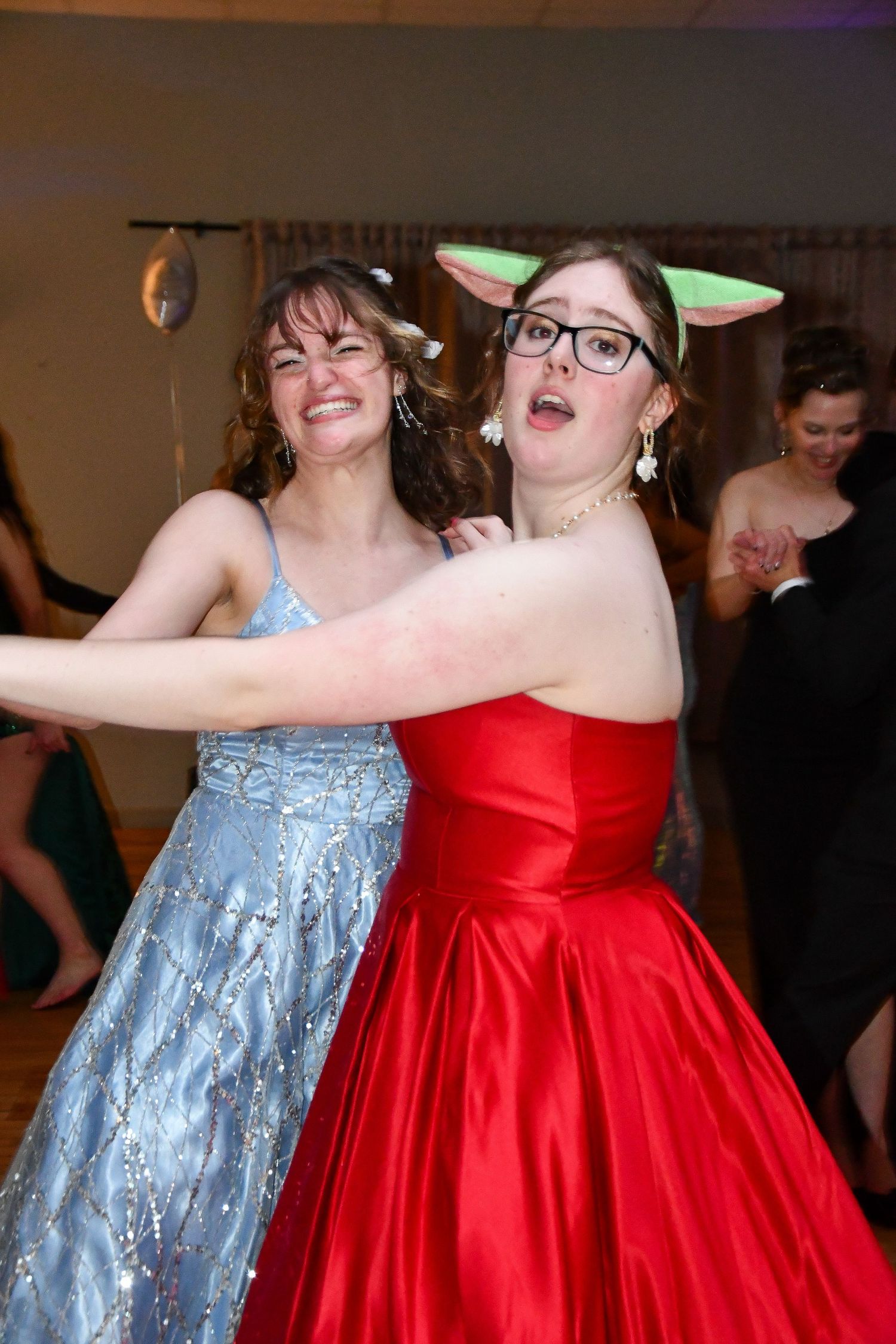 Two friends in elegant prom dresses dance joyfully at formal event.