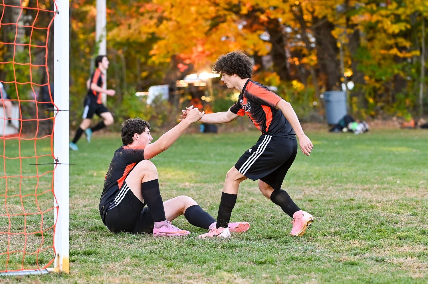 Soccer goalkeeper makes a diving save on grass field during autumn with colorful fall foliage in background.