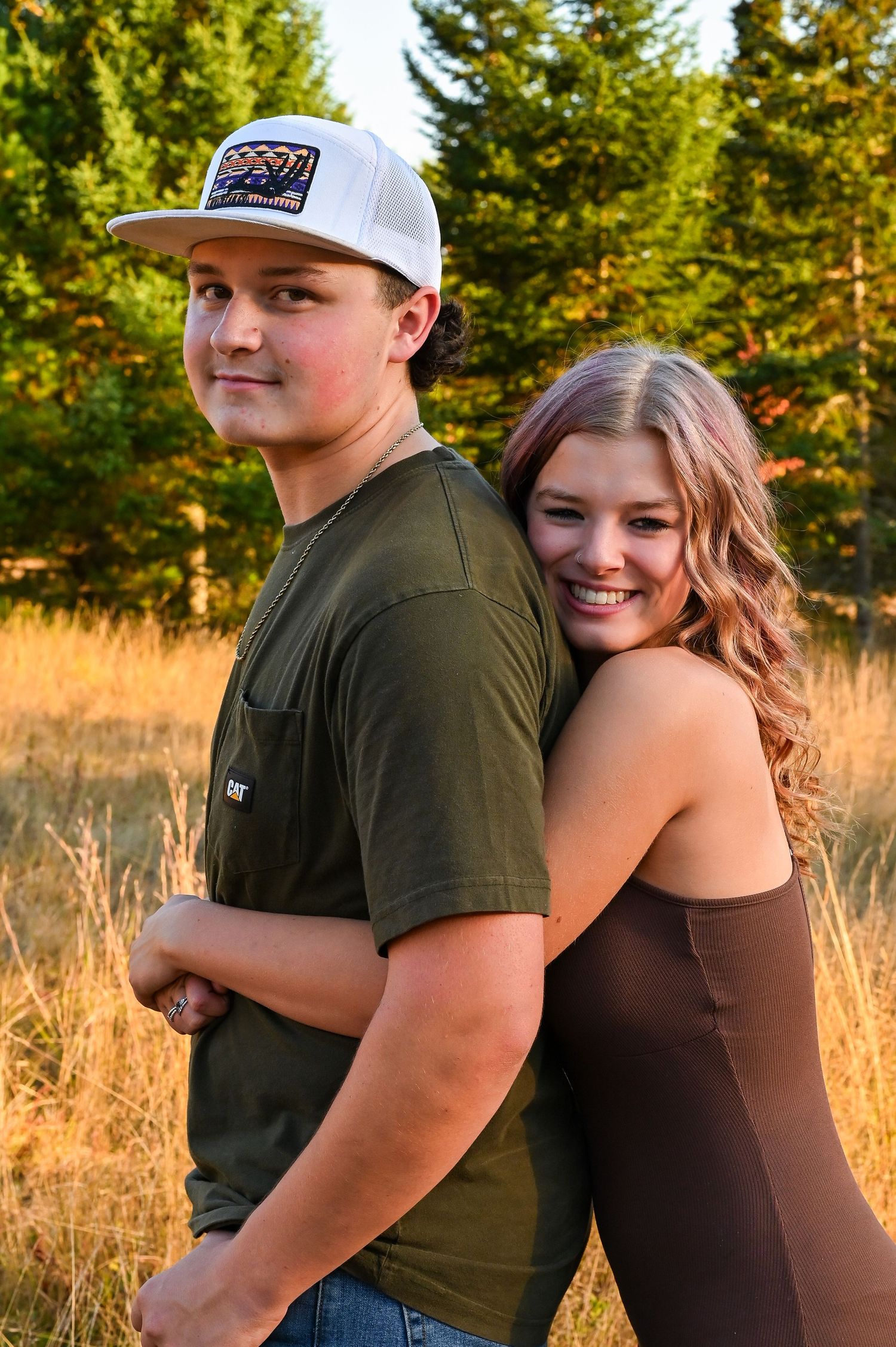 A young couple standing together in a forest, the woman with long brown hair and the man wearing a baseball cap.