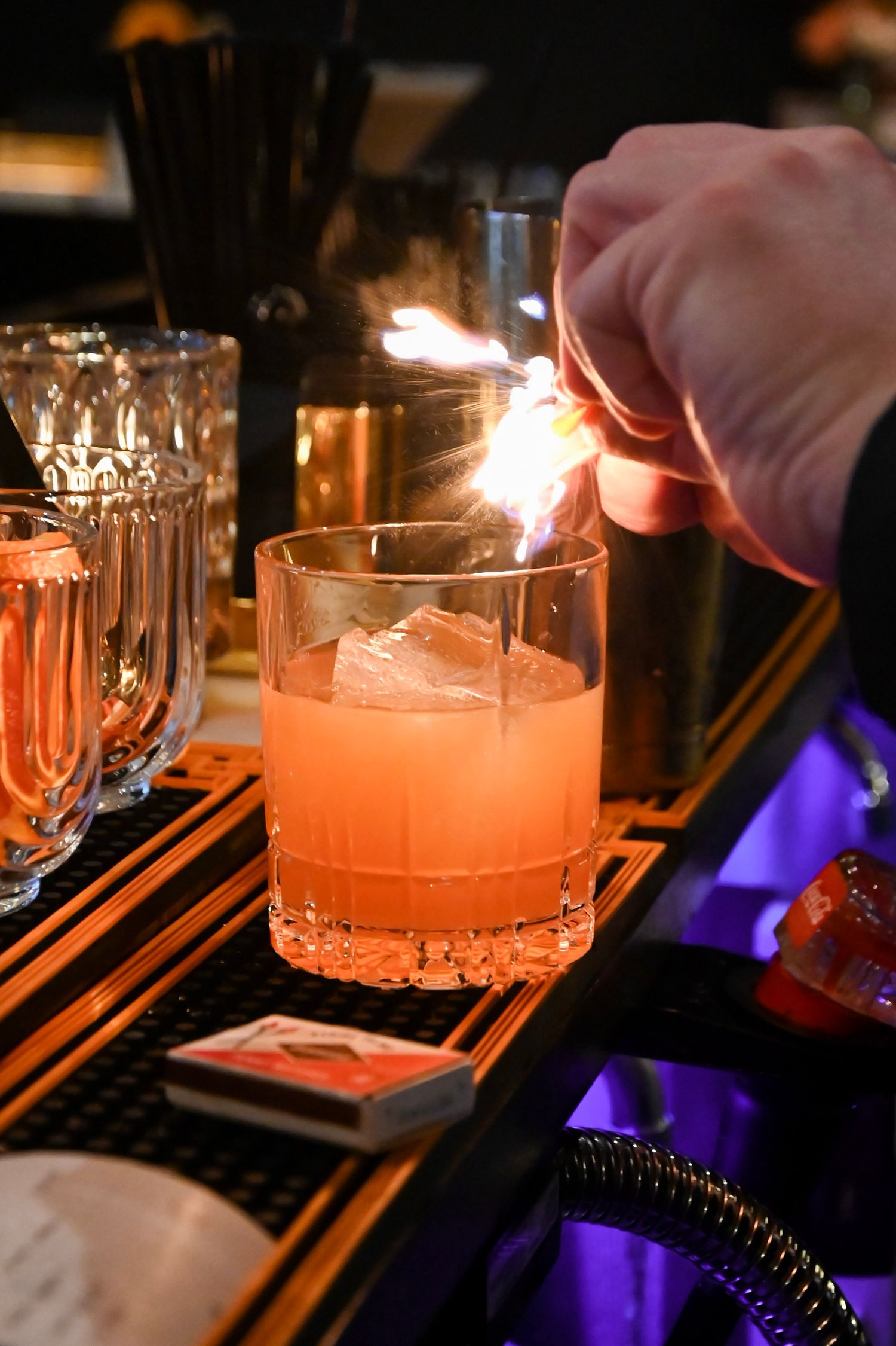 A bartender lights a rose-colored cocktail with fire, creating a dramatic presentation behind the bar at night.