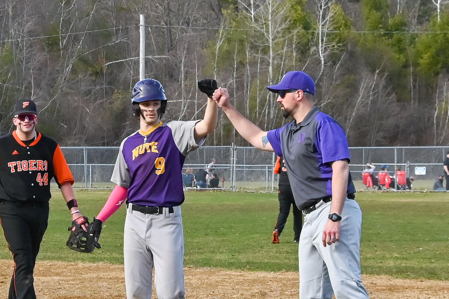 Baseball players and umpire interact on the field during a spring game with trees in the background.