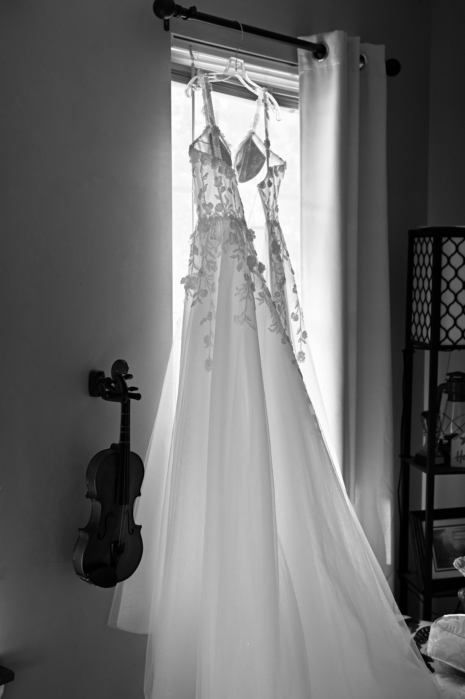 Black and white photo of a lace wedding gown hanging in a window with a violin leaning against the wall nearby.