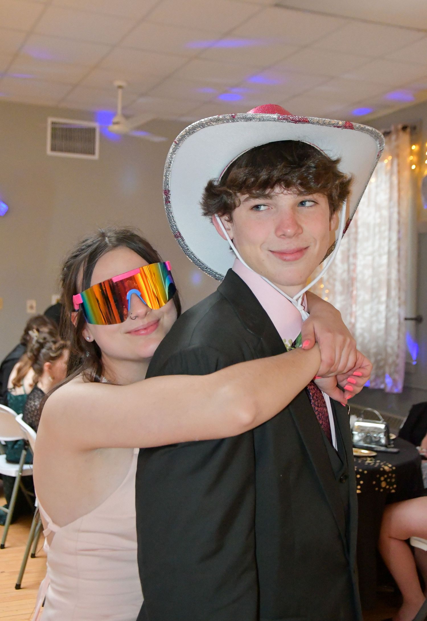 Couple in formal attire posing at a party, one wearing a cowboy hat and the other colorful ski goggles.