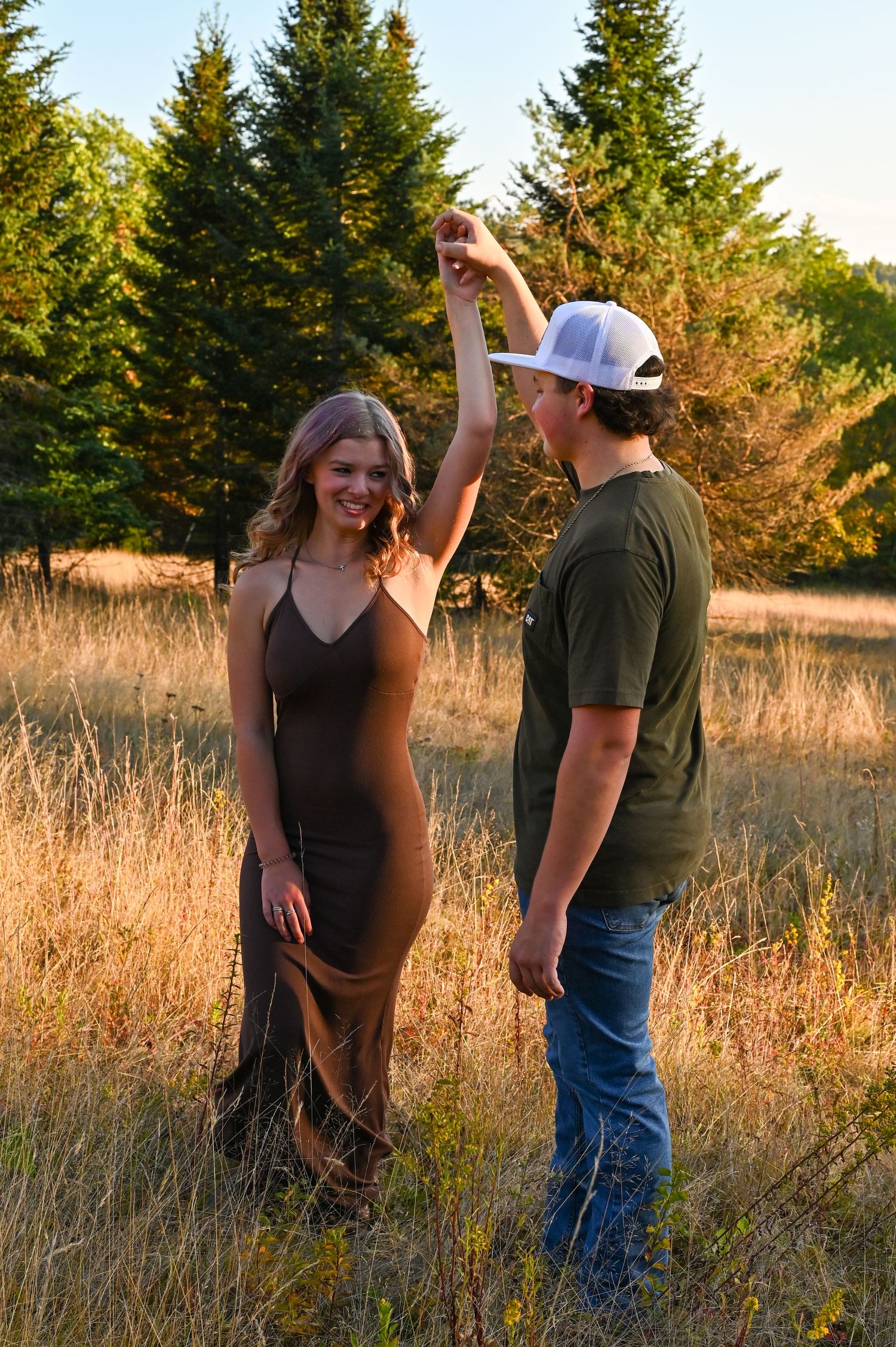 A couple shares a romantic moment in a golden field at sunset with evergreen trees in the background.