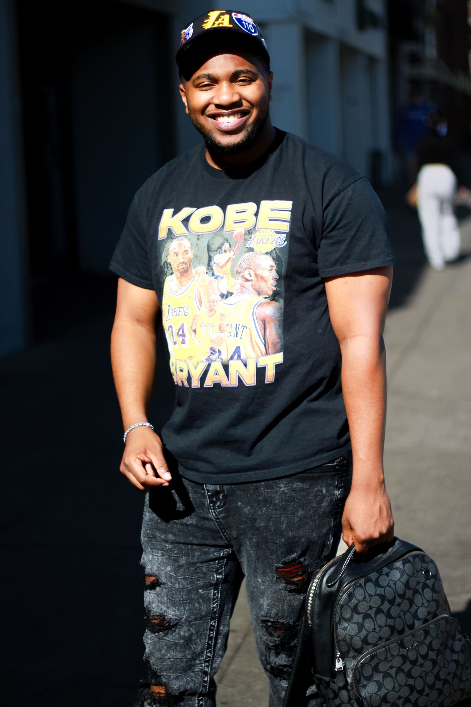 Person wearing Kobe Bryant graphic t-shirt poses with dark backpack in urban setting.