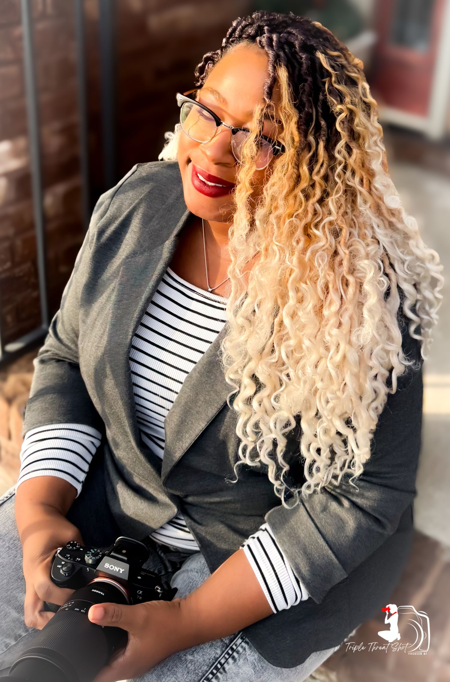Woman with curly ombre hair wearing glasses, striped shirt and blazer smiles while holding a camera.