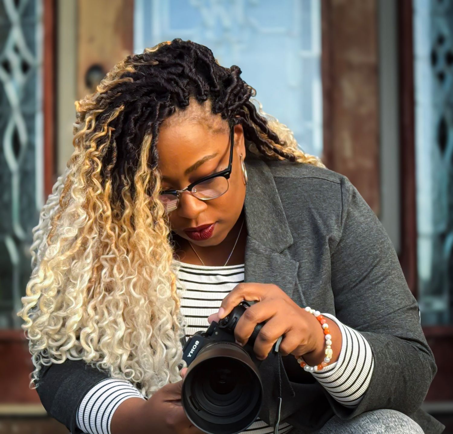 Photographer in striped shirt and gray blazer examines professional camera lens while sitting outside.