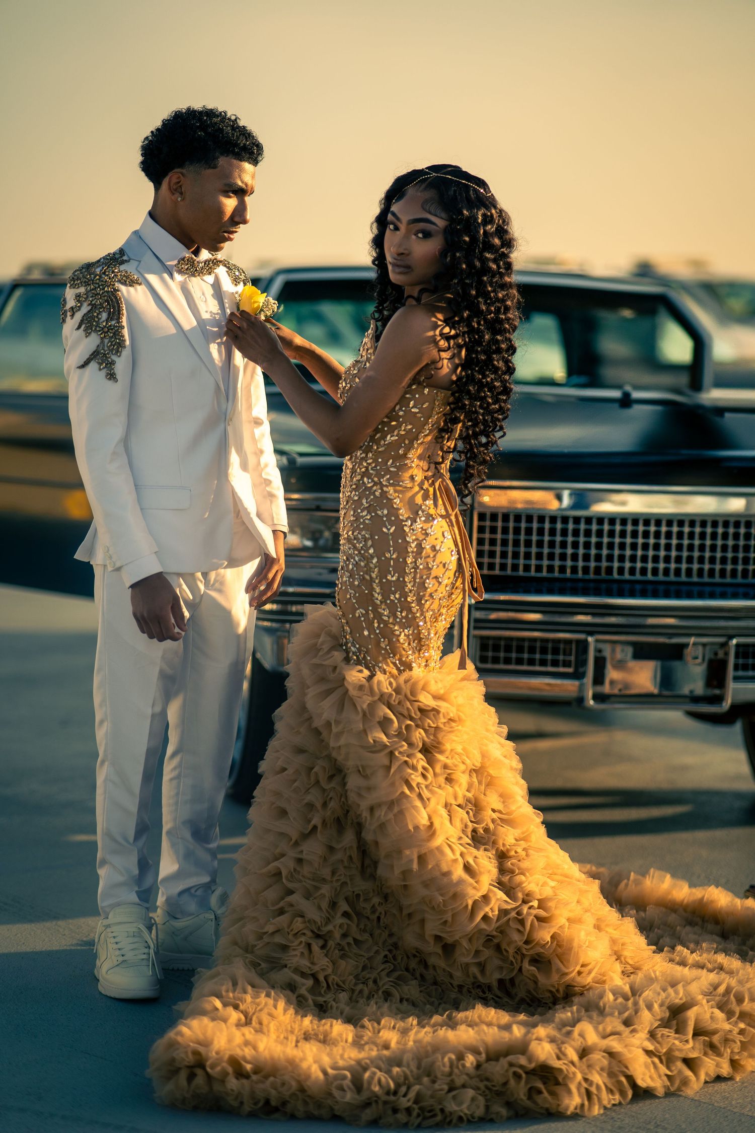 A golden formal dress and white tuxedo shine in sunset light next to a vintage car during a glamorous photoshoot.