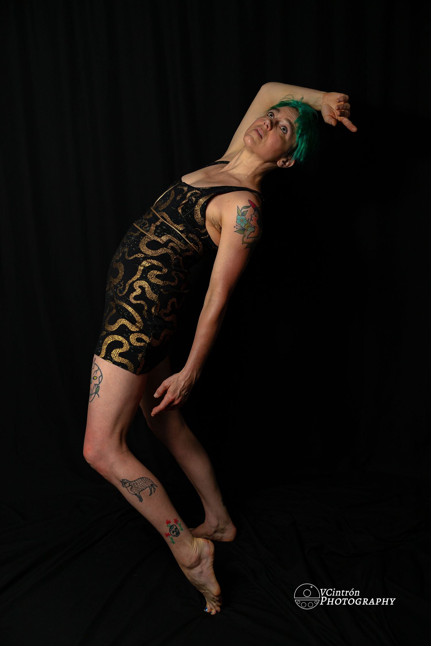 Dancer in black and gold patterned bodysuit with green hair leaps gracefully against dark background in artistic pose.